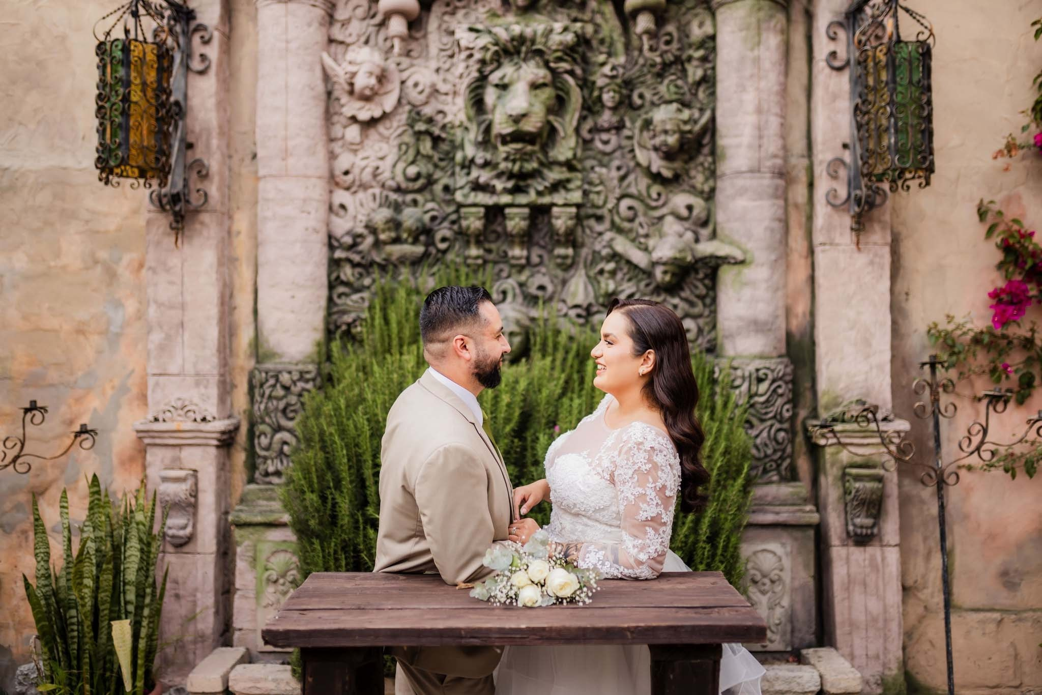 Sesión de fotos de novios en Rosarito Baja California, pareja sentada frente a fuente en Jardín Casa Baga, fotógrafo de bodas en Tijuana con paquetes de fotografía profesional, sesiones románticas y naturales