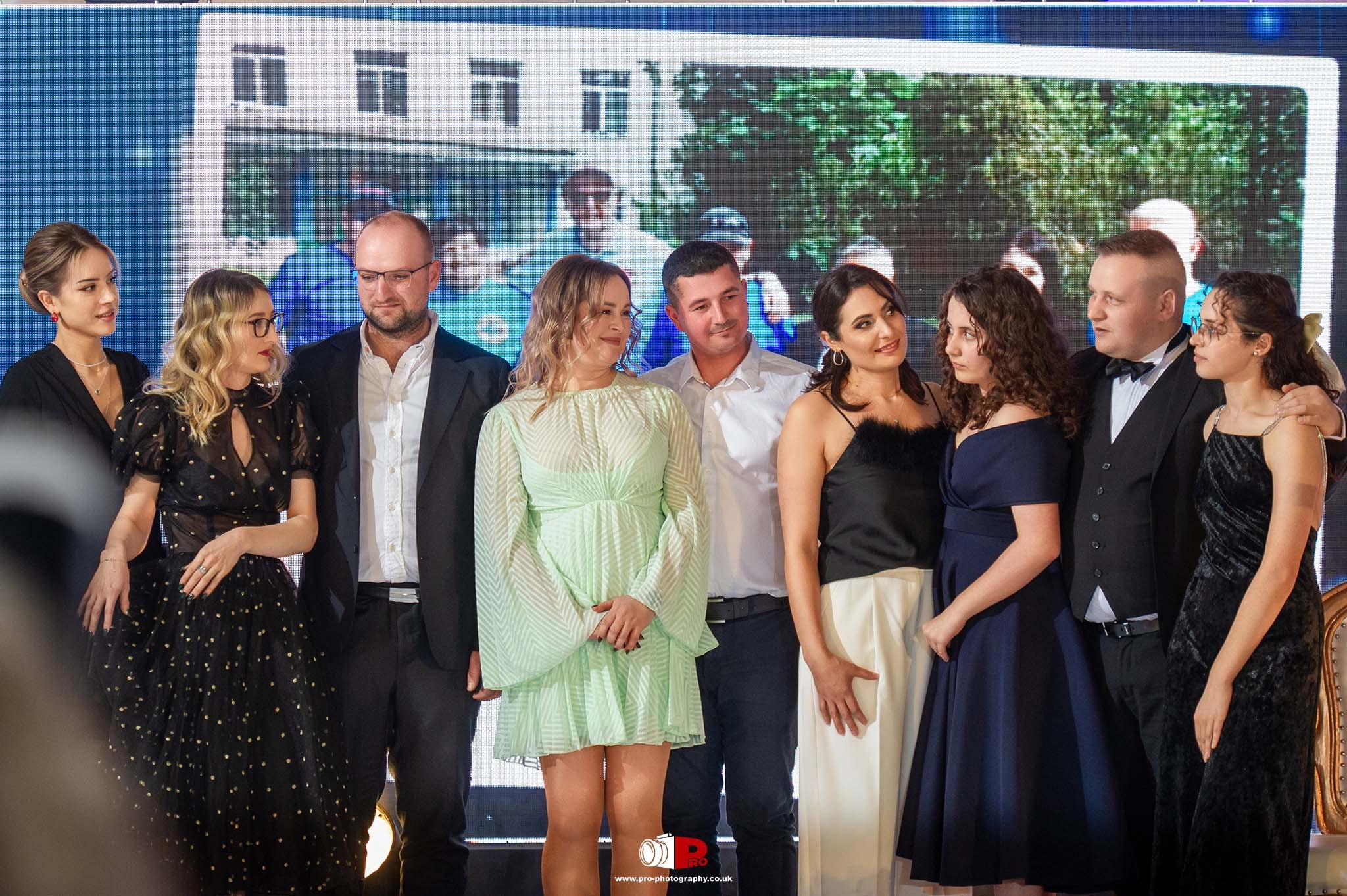 A group of individuals on stage at an awards ceremony with a heartfelt group moment, featuring a large screen backdrop displaying memorable moments.