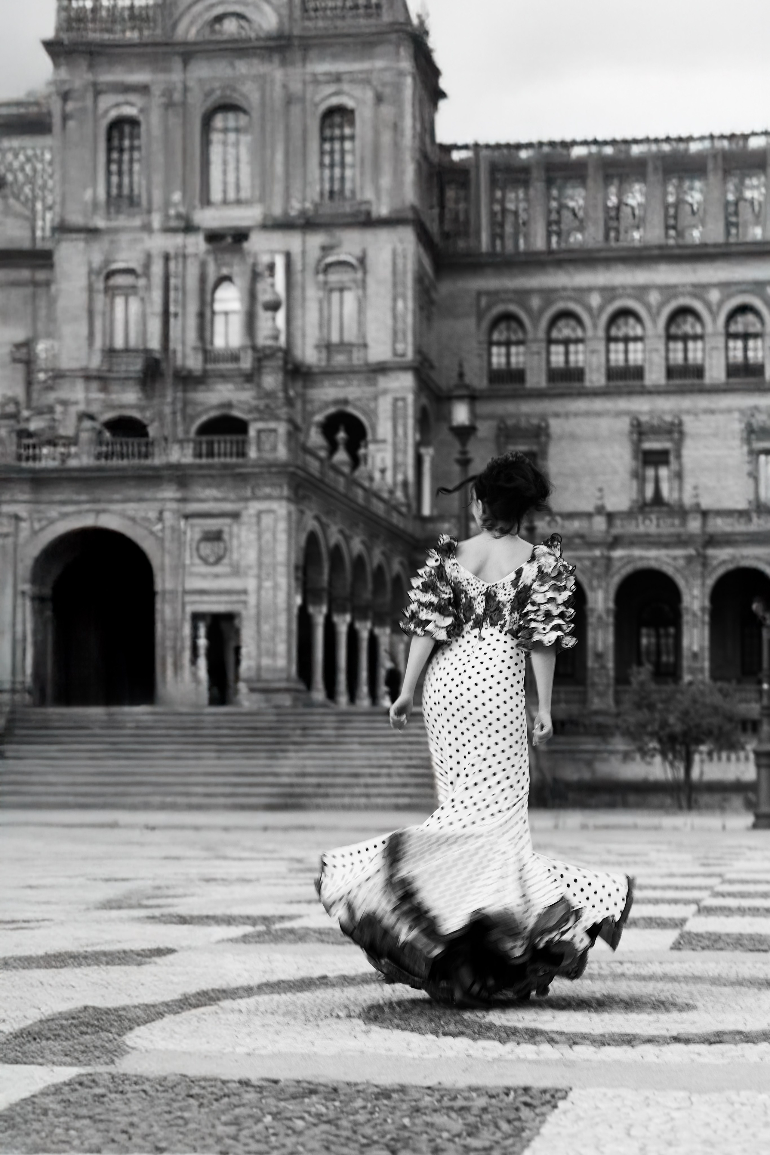 Чёрно-белая портретная фотосессия в Севилье, Испания — женщина в платье фламенко в горошек идёт по площади Испании, демонстрируя вечную элегантность и испанское культурное наследие, идеально для художественной и традиционной портретной съёмки в Севилье.
