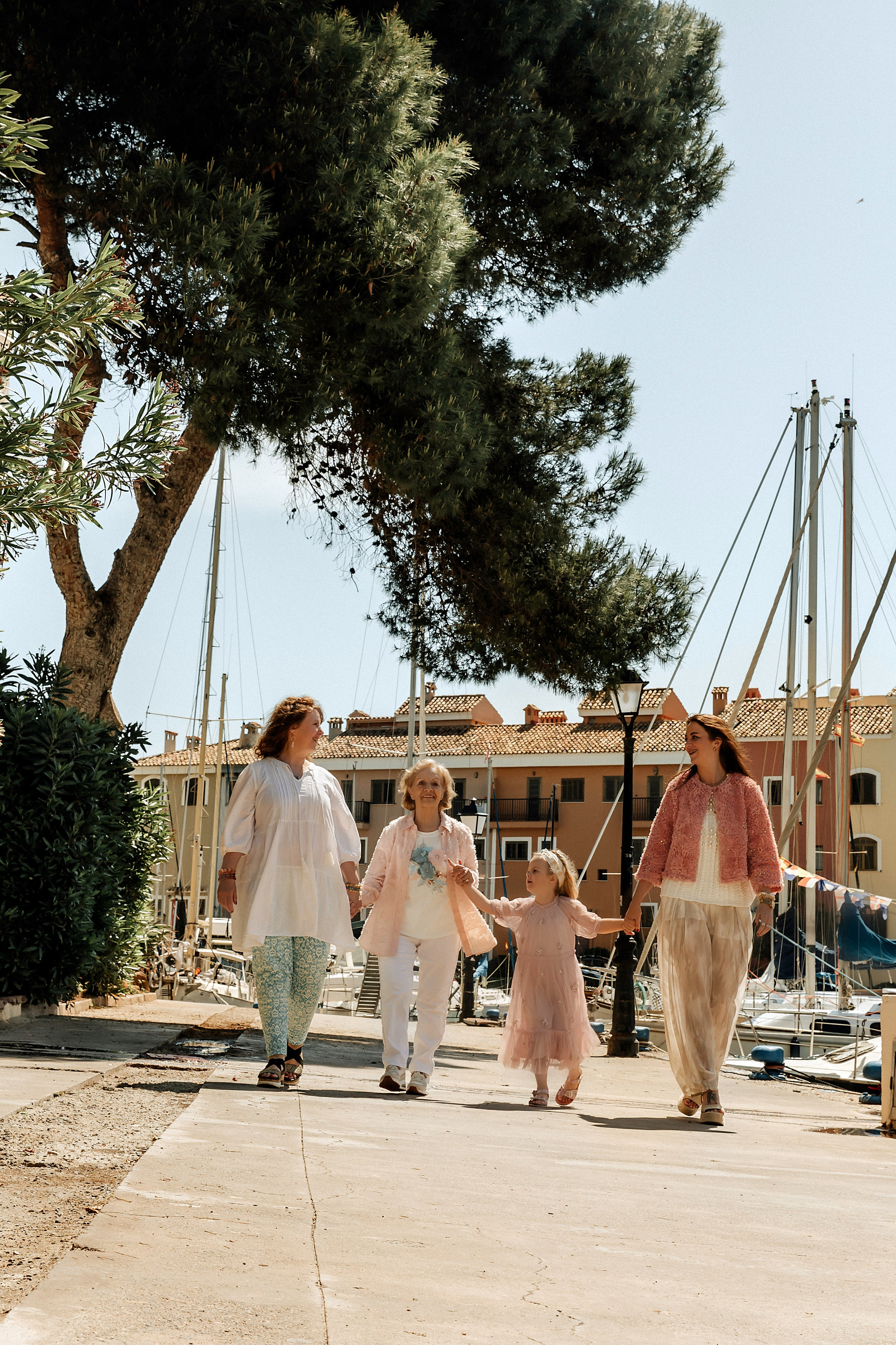 Retrato generacional alegre de cuatro mujeres caminando de la mano cerca del puerto en Port Saplaya, Valencia, España — momento soleado de conexión familiar junto al mar.