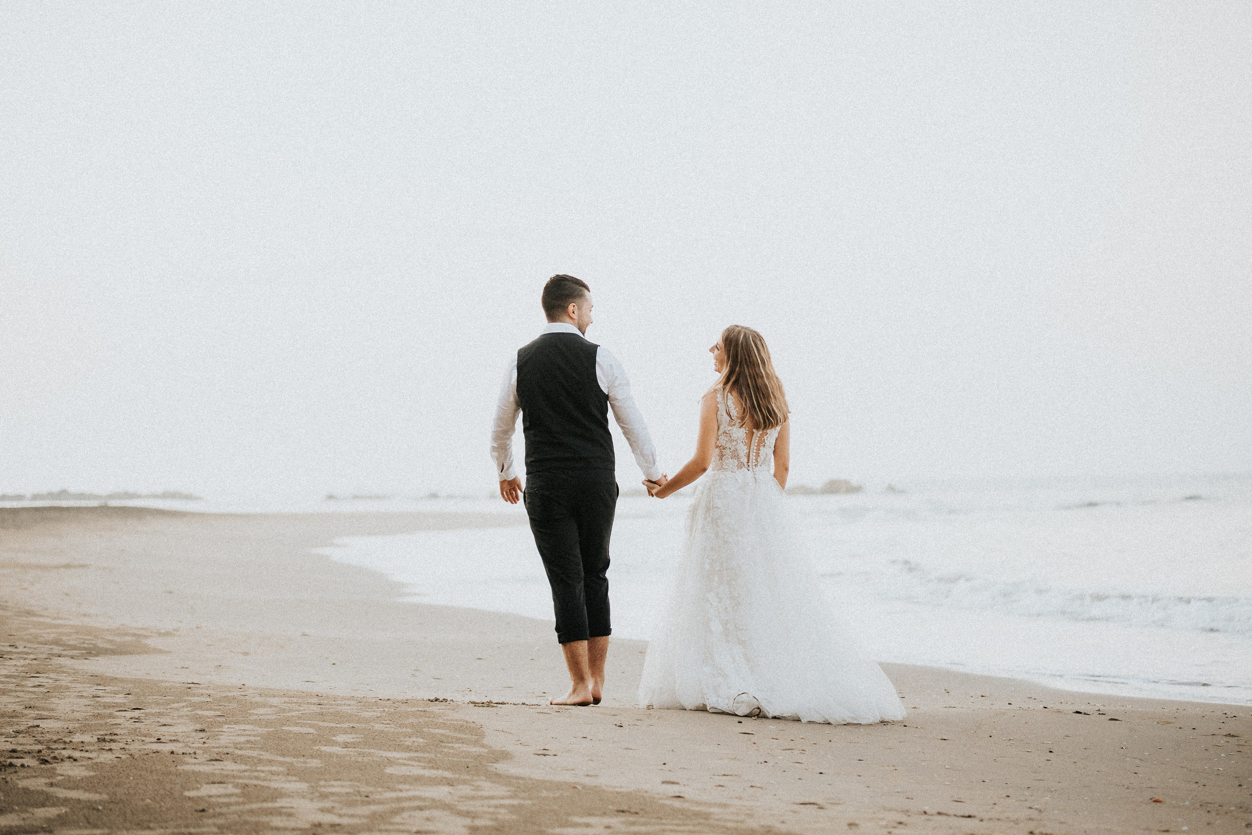 Sposi di spalle si tengono per mano guardando verso l'orizzonte marino, immortale momento di felicità catturato da un fotografo di matrimonio professionista.