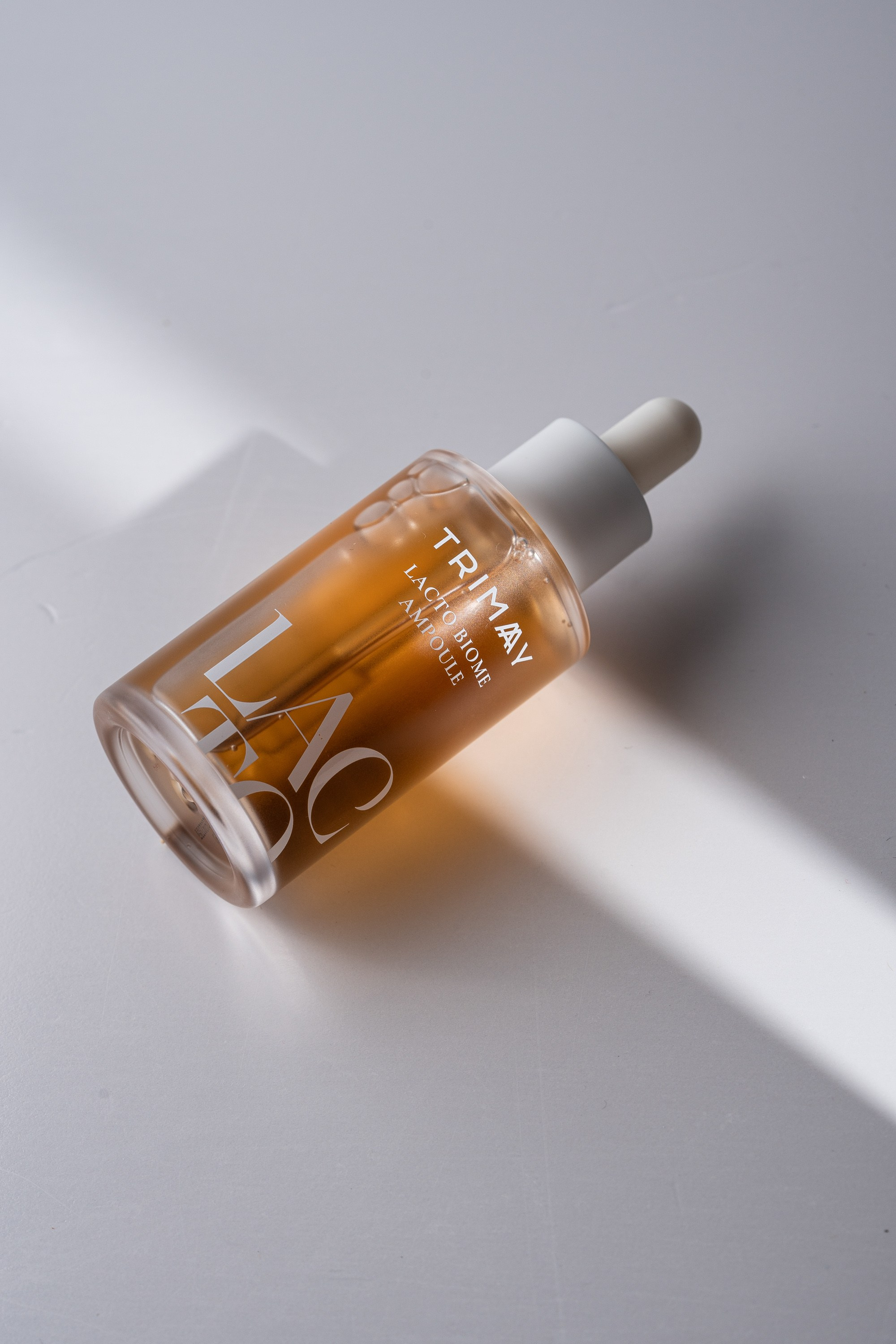 Trimay Lacto Biome Ampoule şüşəsi, ağ fonda işıq şüası effekti ilə minimalistik məhsul fotoşəkili.