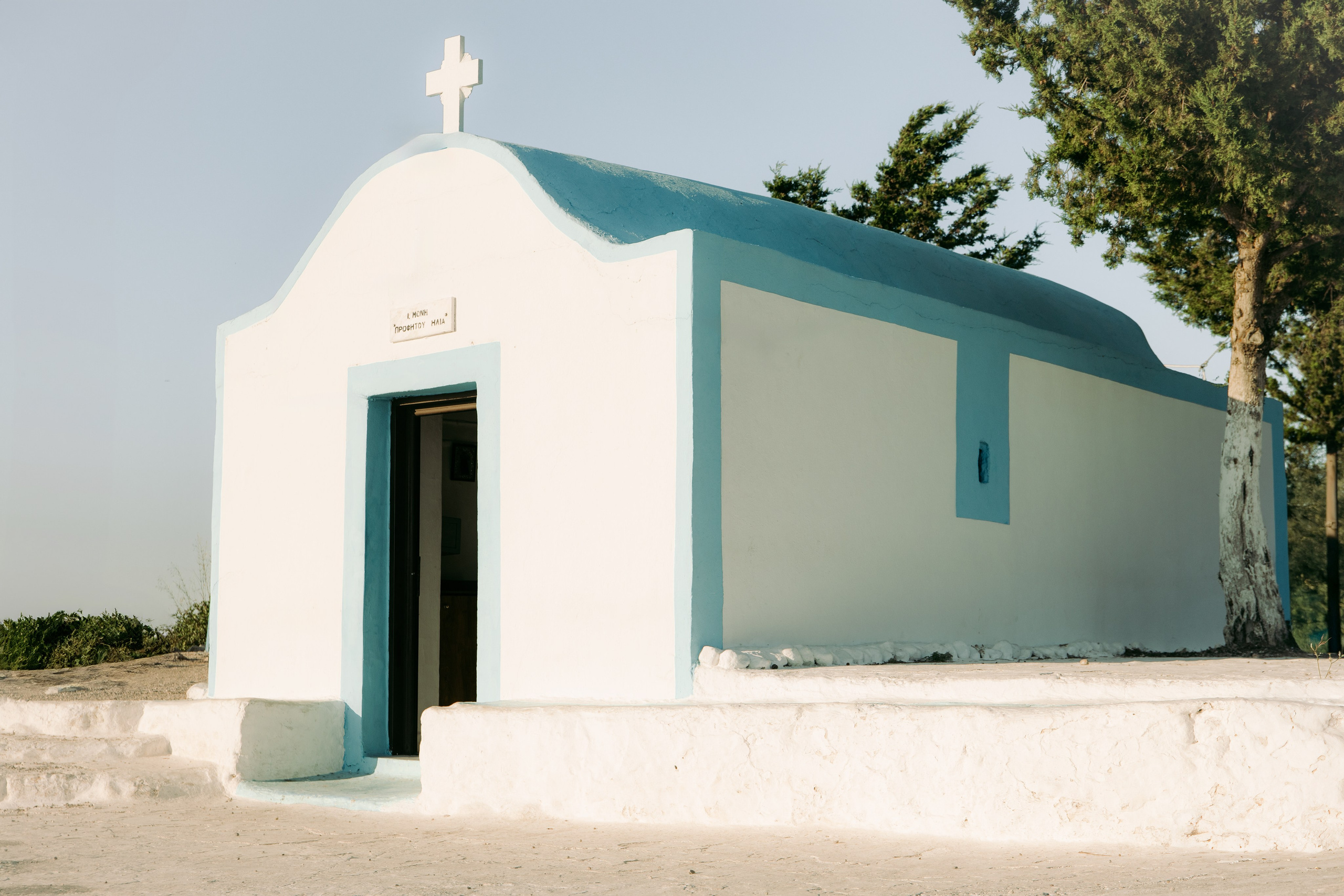 Nahaufnahme der Kapelle Profitis Ilias: Weiße Kalkwände, eine tiefblaue Holztür und ein einfaches Eisenkreuz auf dem Dach. Die Kapelle steht auf einem felsigen Plateau hoch über der Küste von Rhodos."