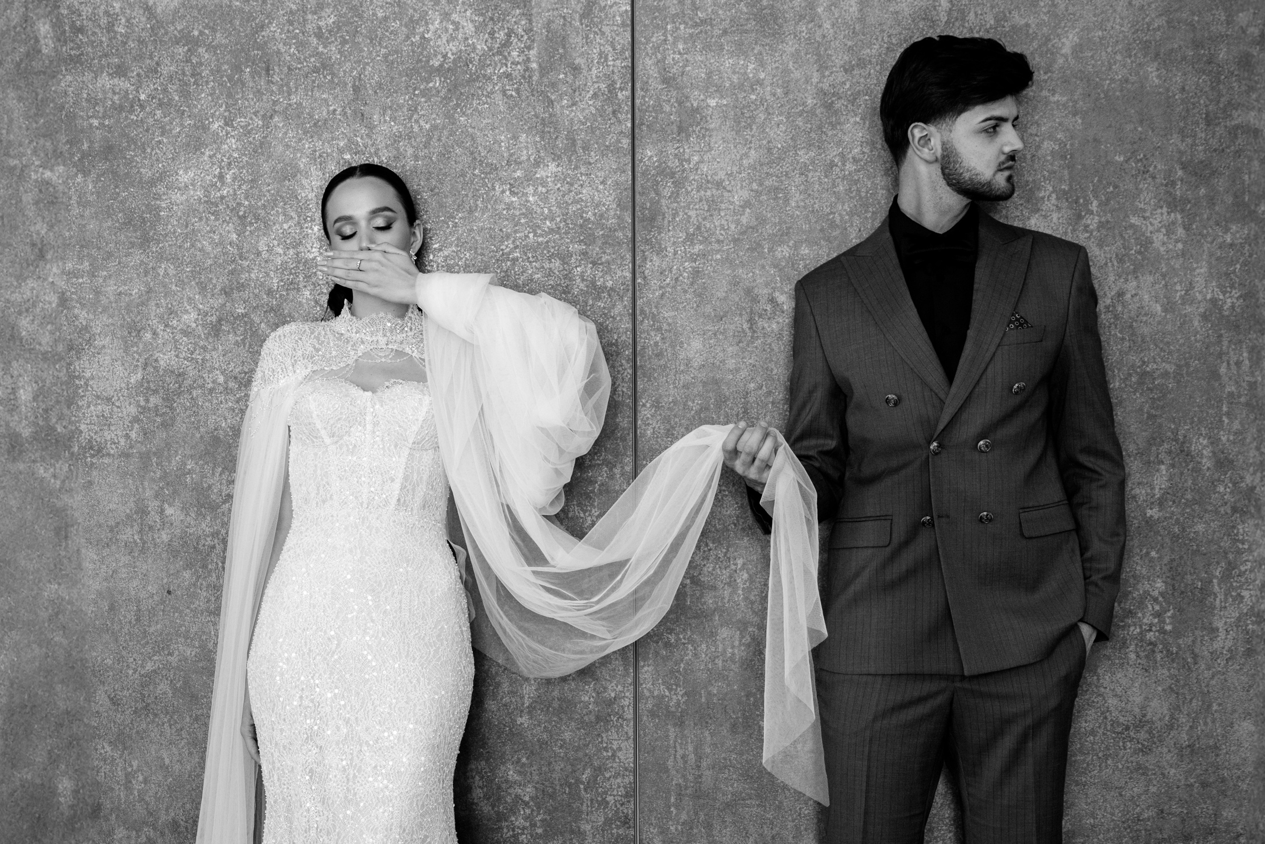 Cuplu în ținută formală, ținându-se de mână, în fața unui perete texturat