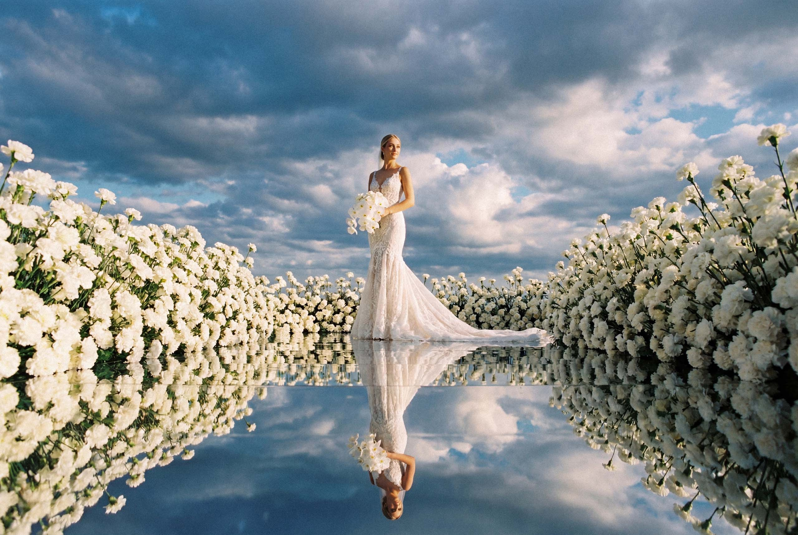 Luxury wedding decor with a mirrored aisle: a bride stands surrounded by an installation of white carnations under a dramatic cloudy sky.