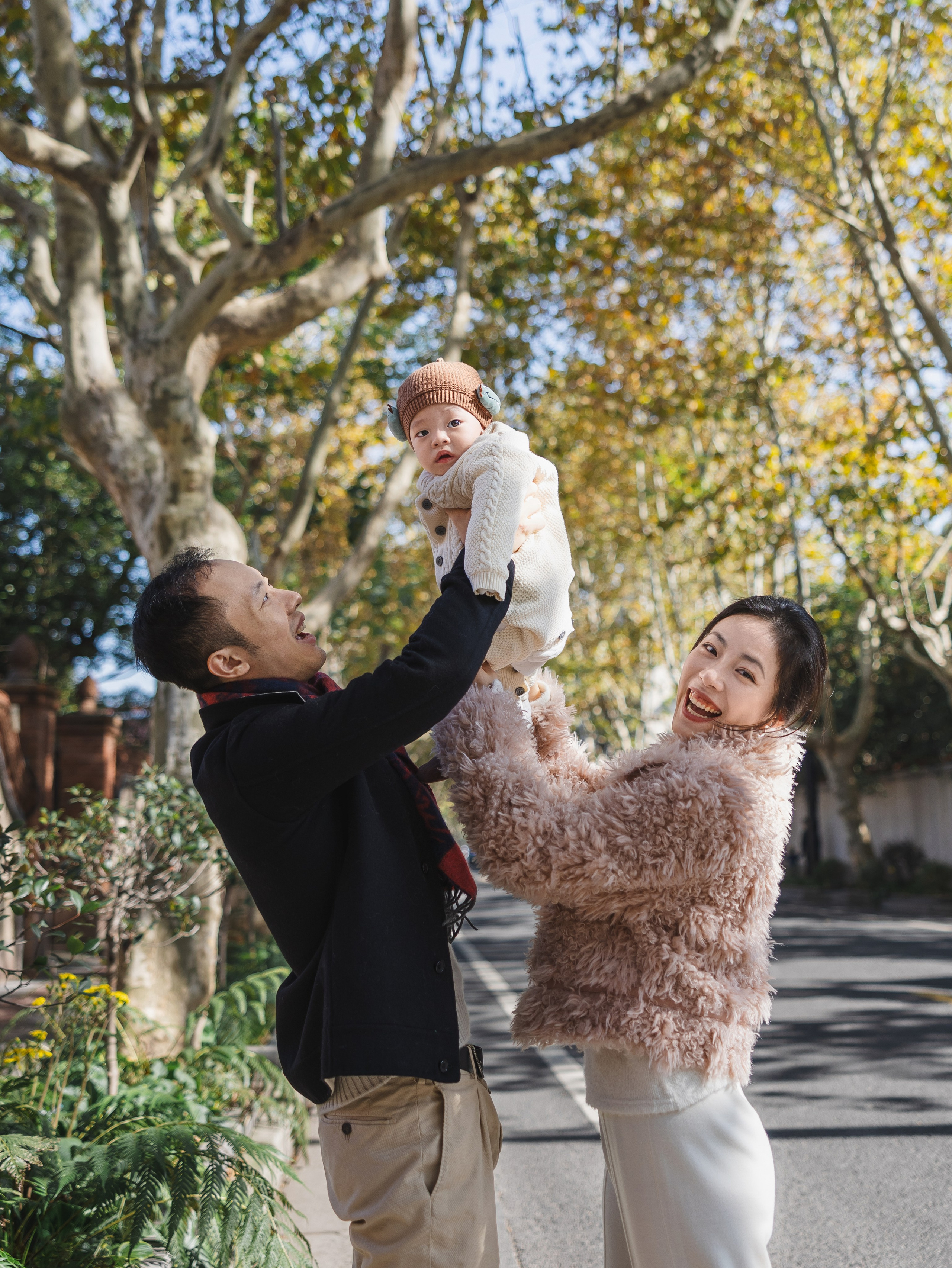 In the late autumn of Shanghai, the parents lift their little baby high, with warm yellow leaves in the background.