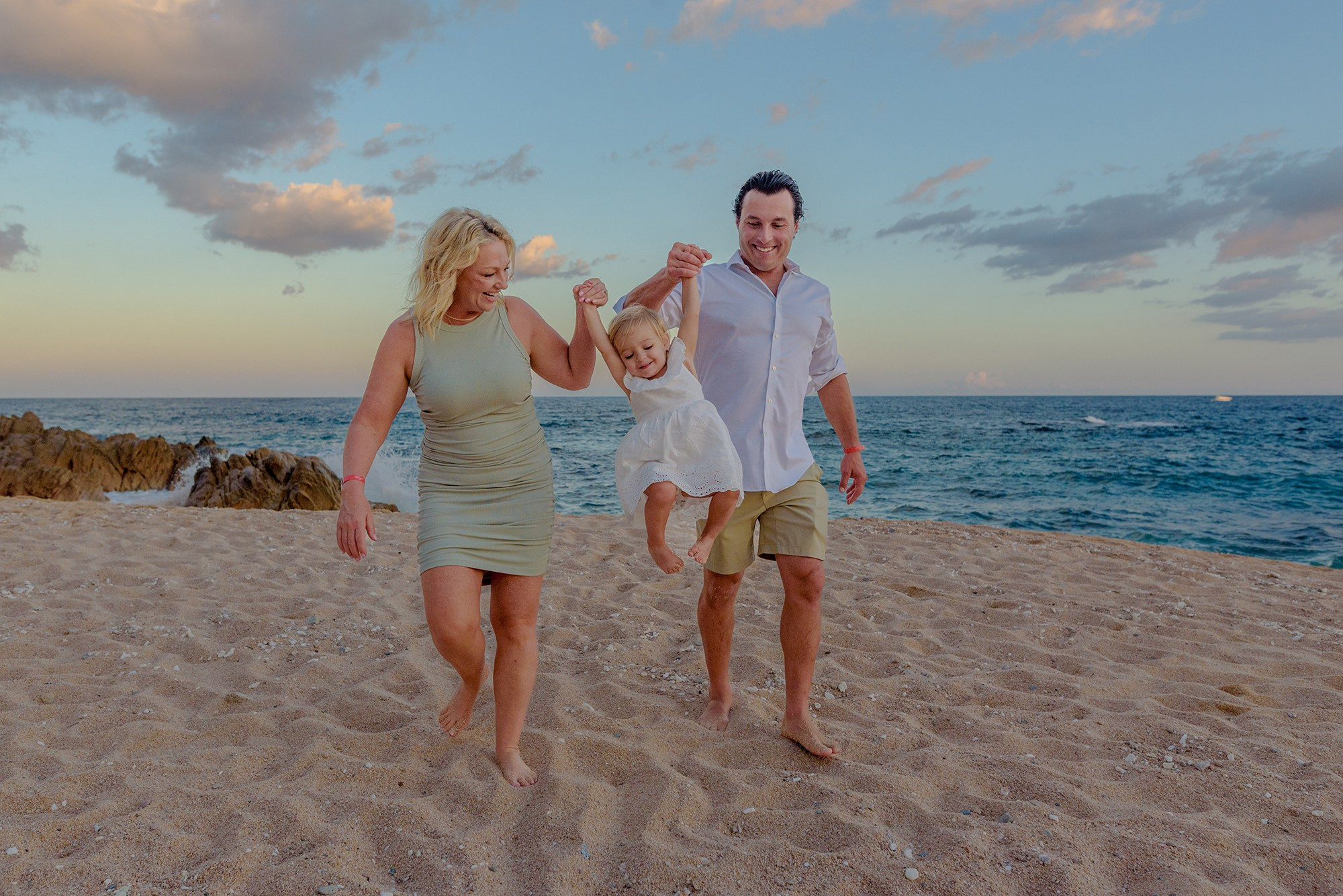 Family beach session in Los Cabos – parents swinging their four year old daughter by the hands at sunset, joyful lifestyle family photography