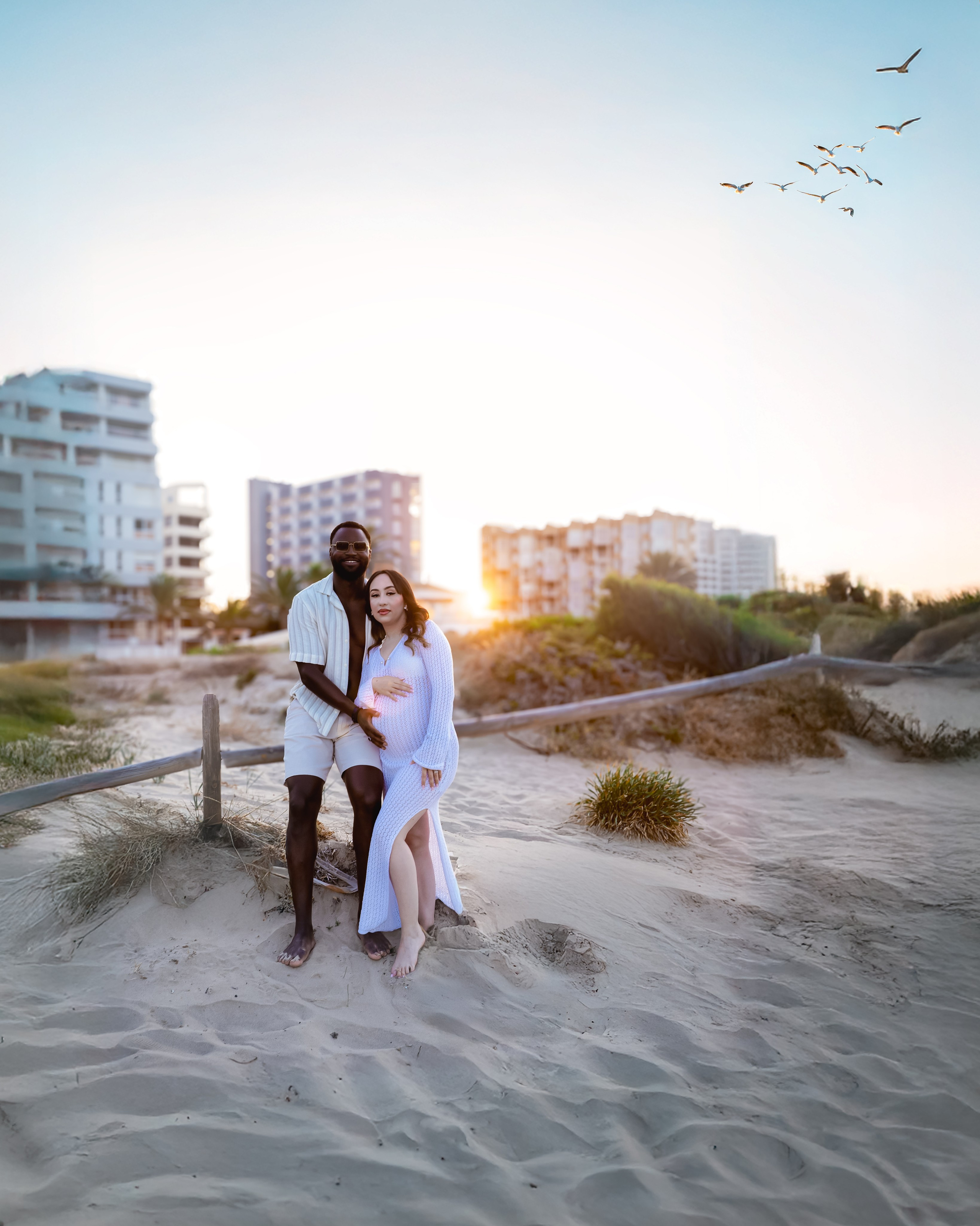 Sesión al atardecer en Calpe, España — pareja embarazada de pie en la playa con edificios al fondo, capturando amor, ilusión y luz cálida. Perfecta para quienes buscan sesiones de embarazo en la playa en Calpe y la costa.