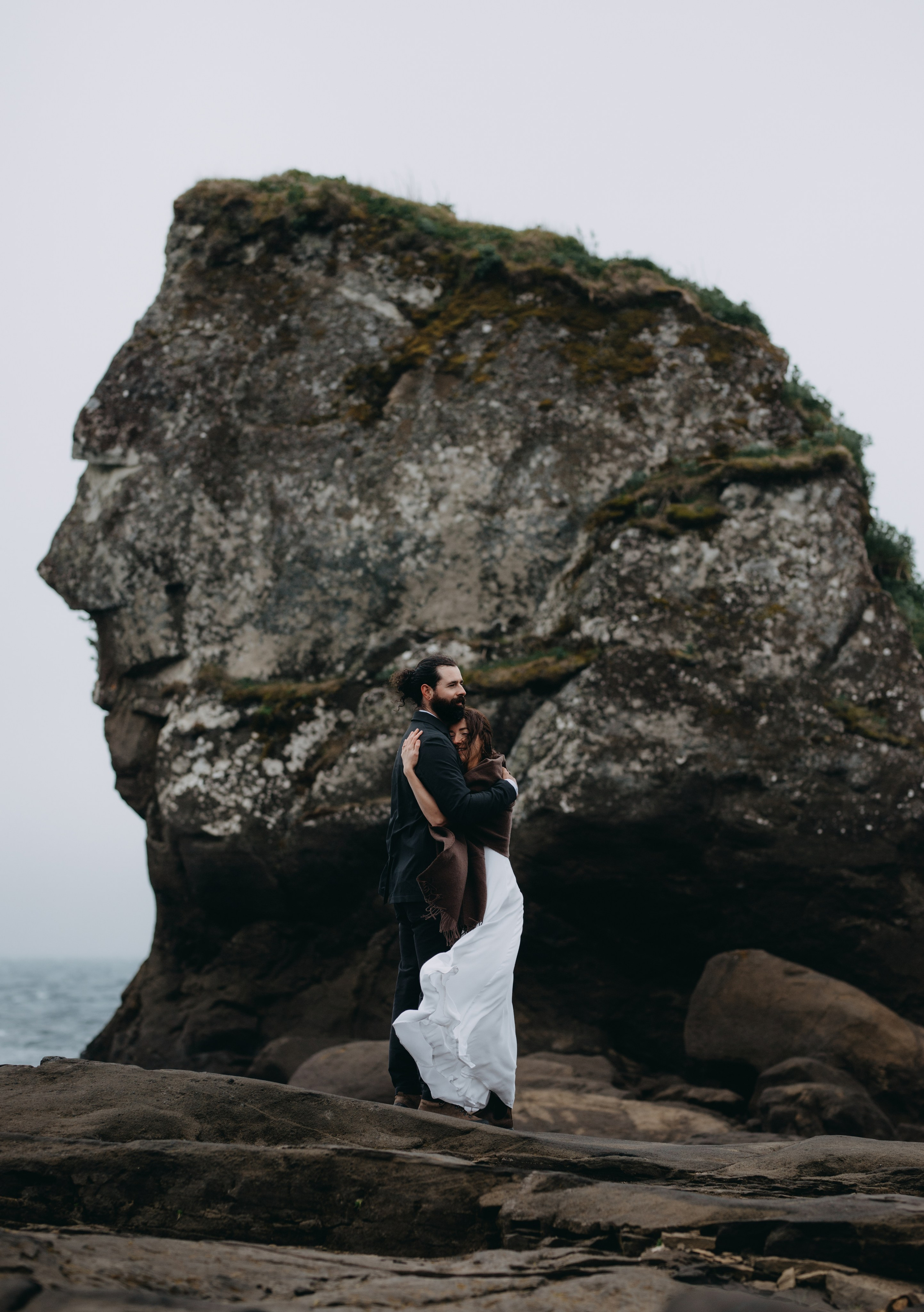 Groom wrapping his arms around the bride as they stand on the rocky edge of Kleifarvatn, braving the cold Icelandic wind.