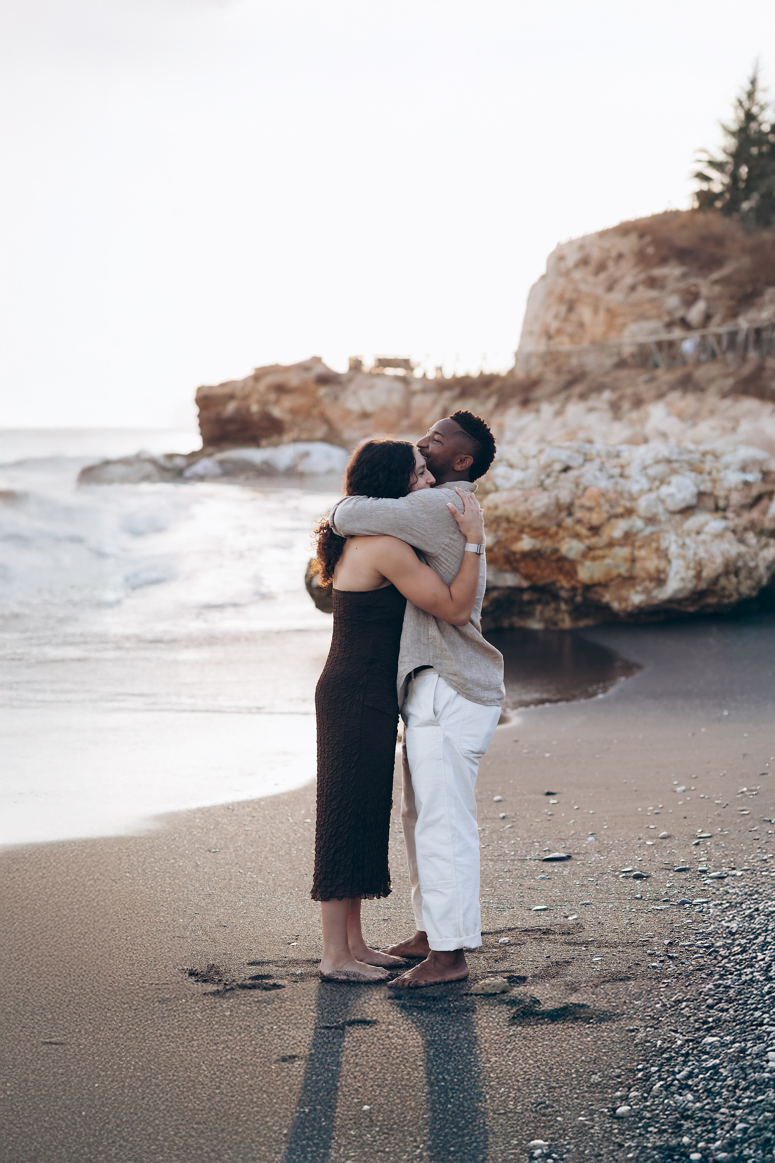 Couple embracing at the water’s edge during a destination engagement session in Málaga. Romantic proposal photography on the Costa del Sol capturing soft light and ocean waves.
