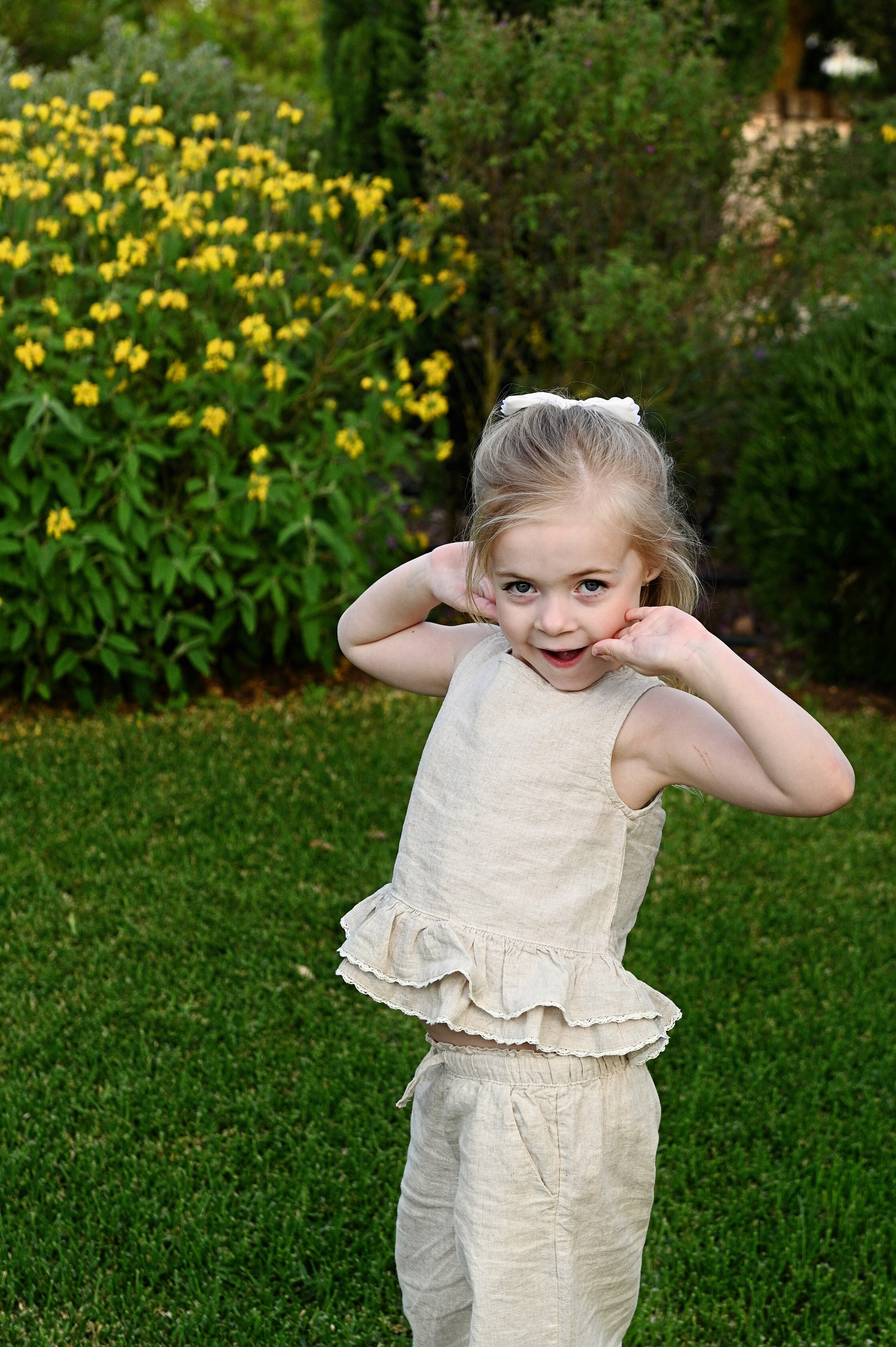 Servizio fotografico per bambini in Puglia: una bambina in un completo di lino crema posa in modo divertente sul prato verde con un cespuglio di fiori gialli sullo sfondo.