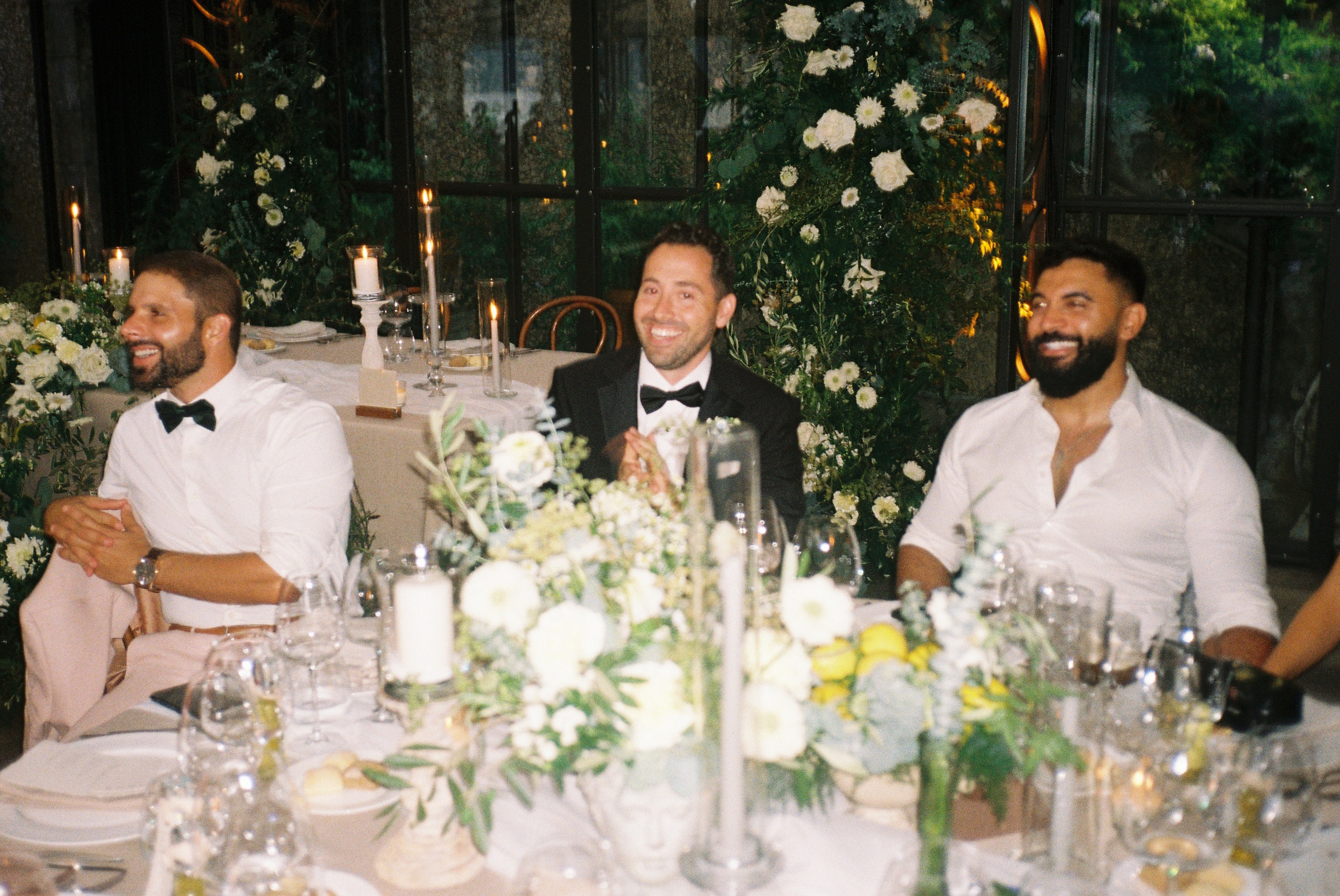 Men in formal attire laugh and enjoy dinner at a beautifully decorated reception table.