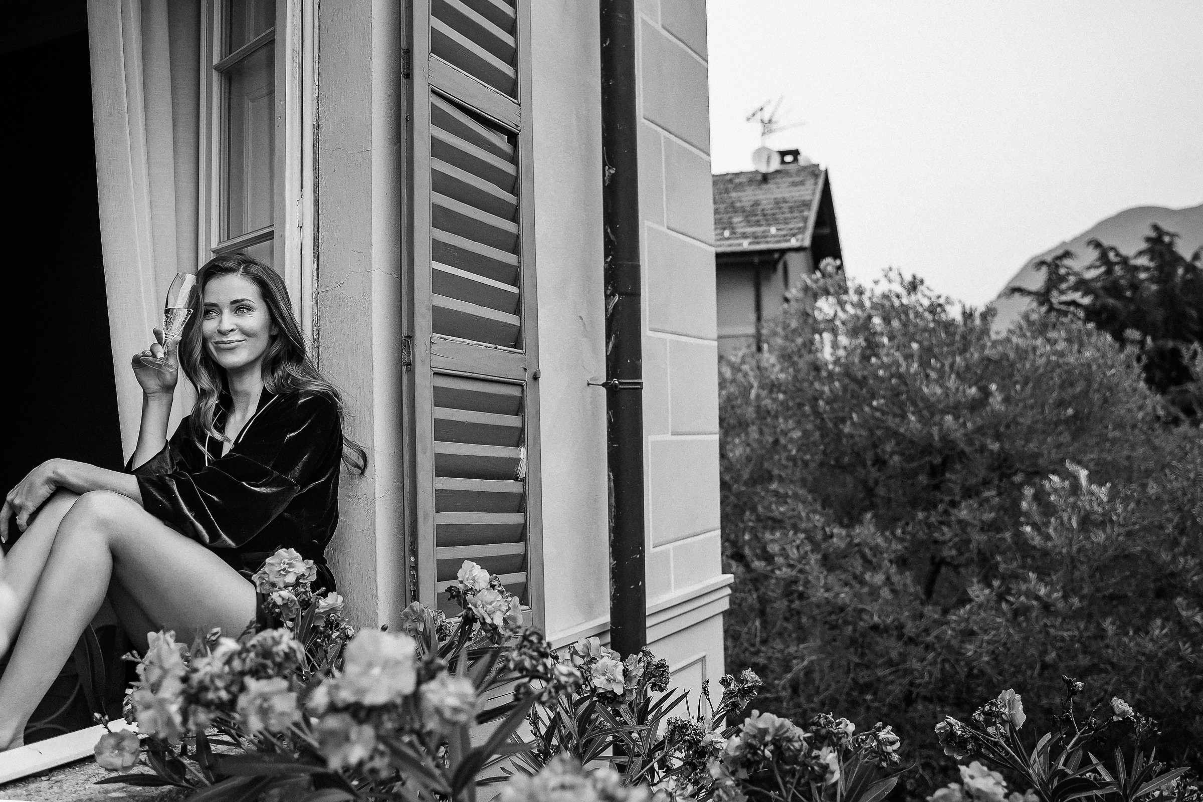 The bride with long hair sits on a windowsill wearing a dark robe, holding a glass in one hand. Shutters frame the window, with blooming flowers in the foreground and a tiled-roof house, trees, and mountain scenery in the background.