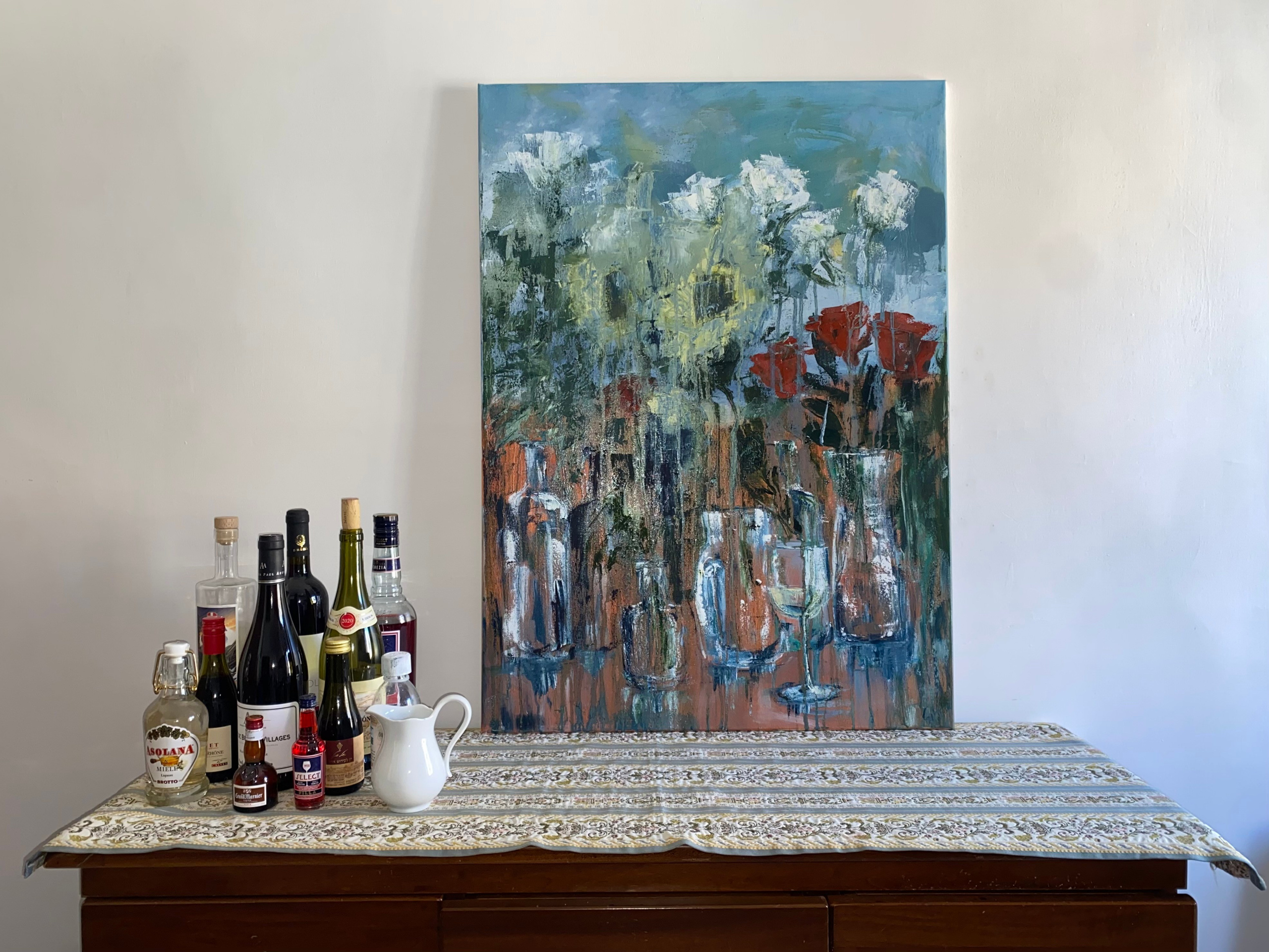 Expressive acrylic still life of a bar table with bottles, wine glasses, and a patterned tablecloth, rendered in warm reds and deep greens. Painting by Ekaterina Solopova addressing rituals of drinking and shared moments in bars restaurants and aperitivo time. With flowers melting