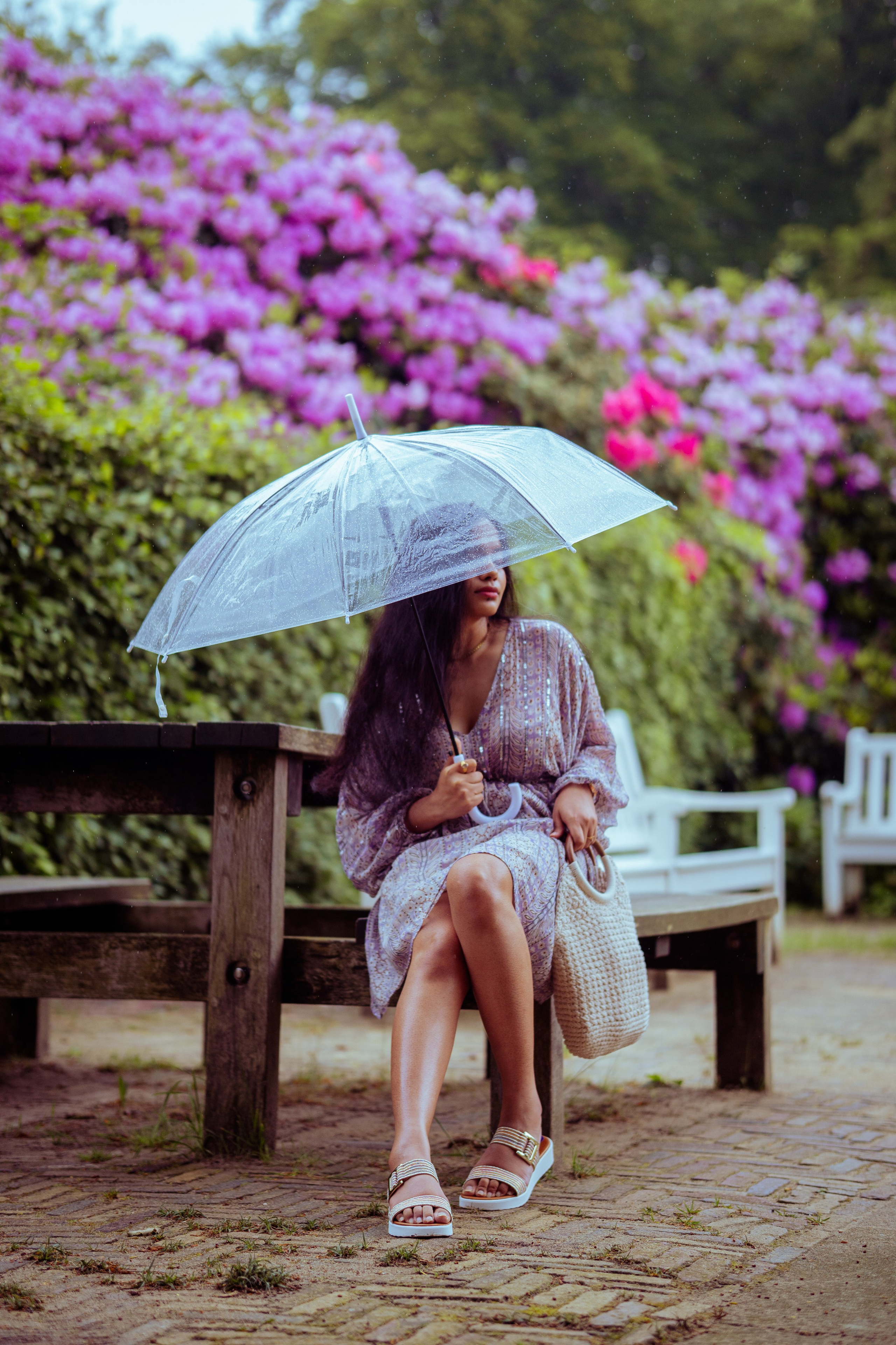 woman sitting on a wooden bench with flowers as background and holding a transparent umbrella