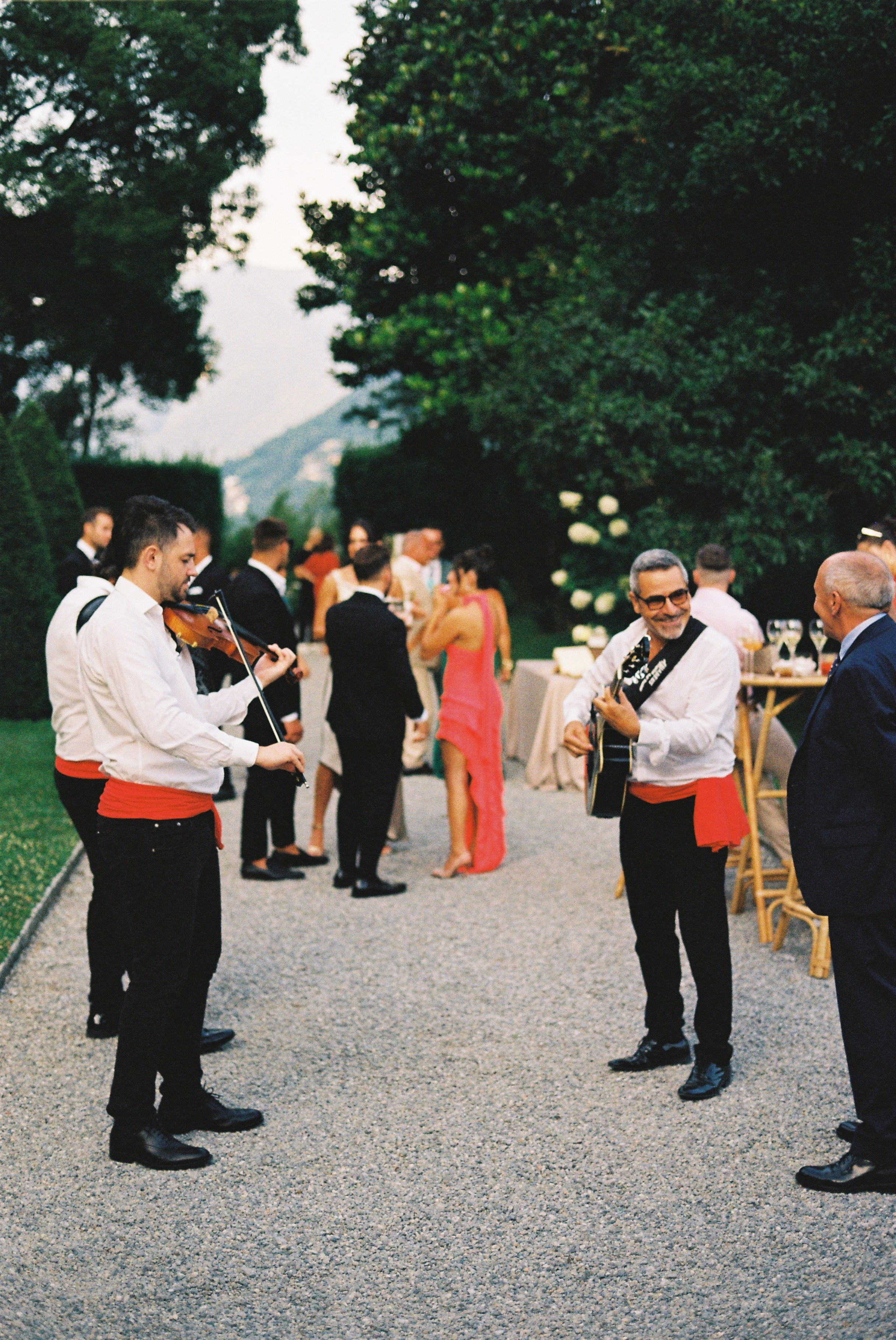 Musicians play violin and guitar as guests mingle during outdoor wedding reception.