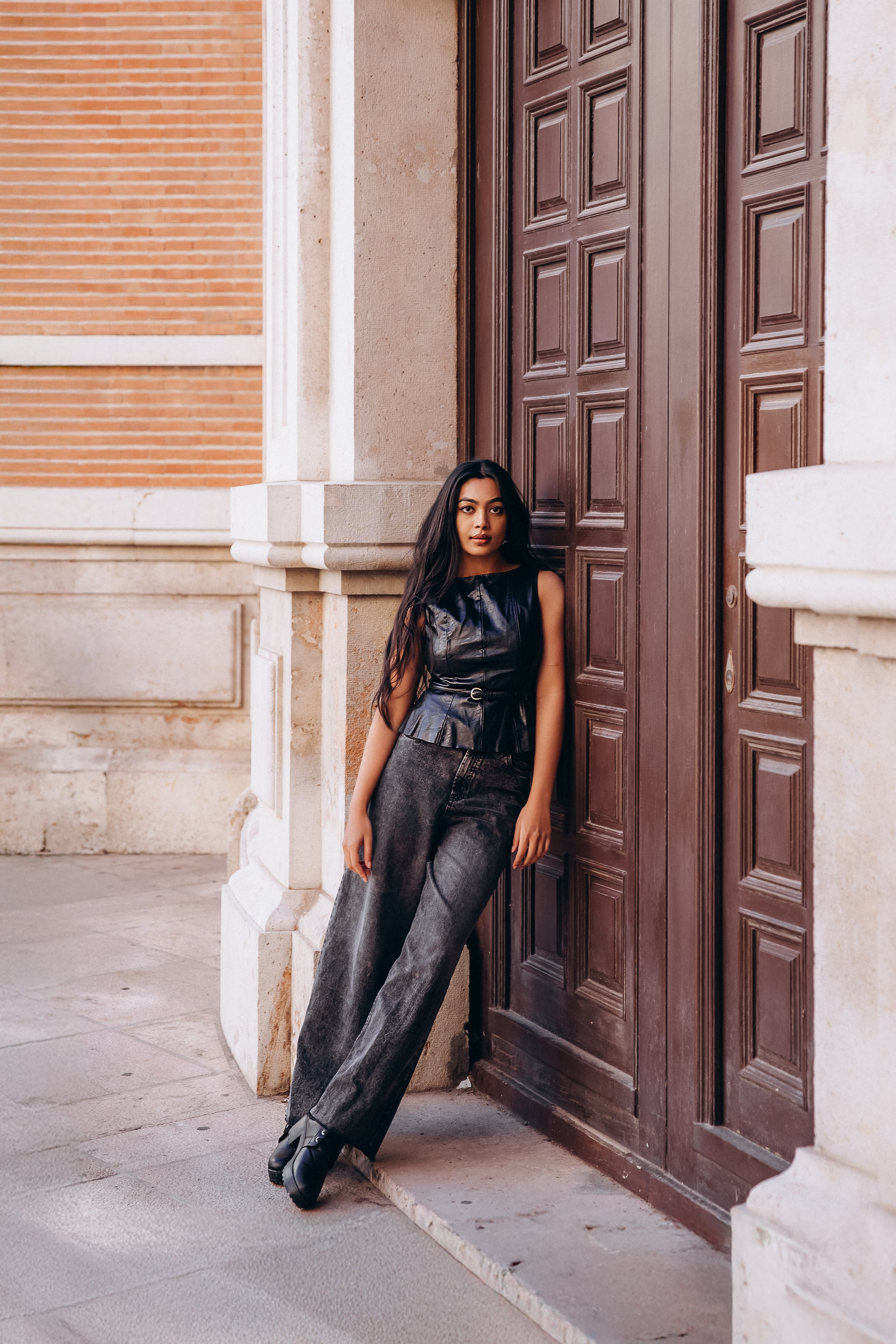 Полноформатный женский портрет, снятый в Валенсии, Испания — уверенная женщина в элегантной черной одежде, прислонившаяся к резной деревянной двери. Идеально подходит для демонстрации стильных и мощных женских портретных фотосессий в Валенсии и по всей Испании.