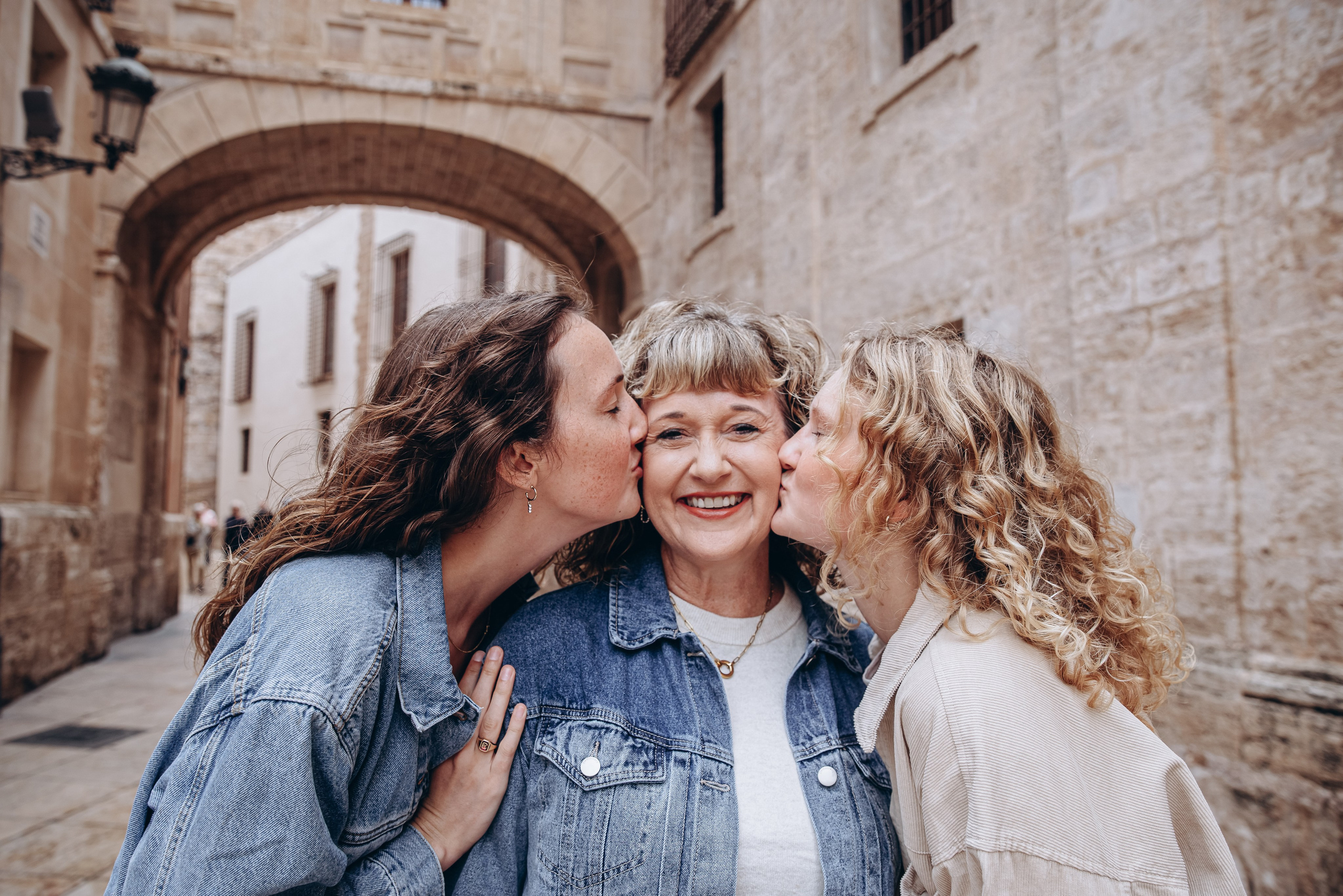 Tres generaciones compartiendo amor durante una sesión familiar en Valencia, España — dos hijas besando a su madre sonriente en un encantador callejón del casco antiguo. Ejemplo perfecto de fotografía familiar emotiva en Valencia para quienes buscan momentos naturales y conmovedores en sus sesiones en España.