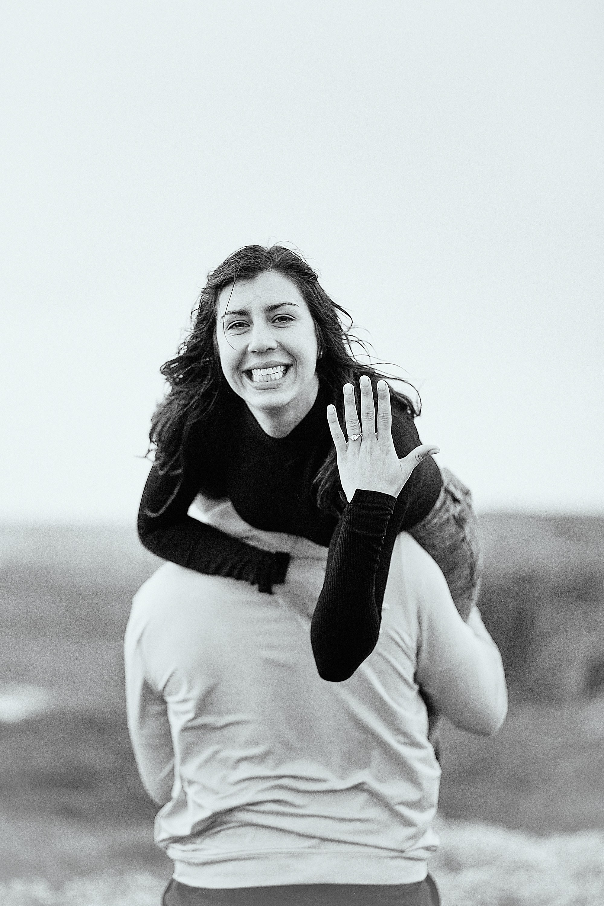 A joyful woman being carried on her partner's back, excitedly showing off her new engagement ring in Ireland.