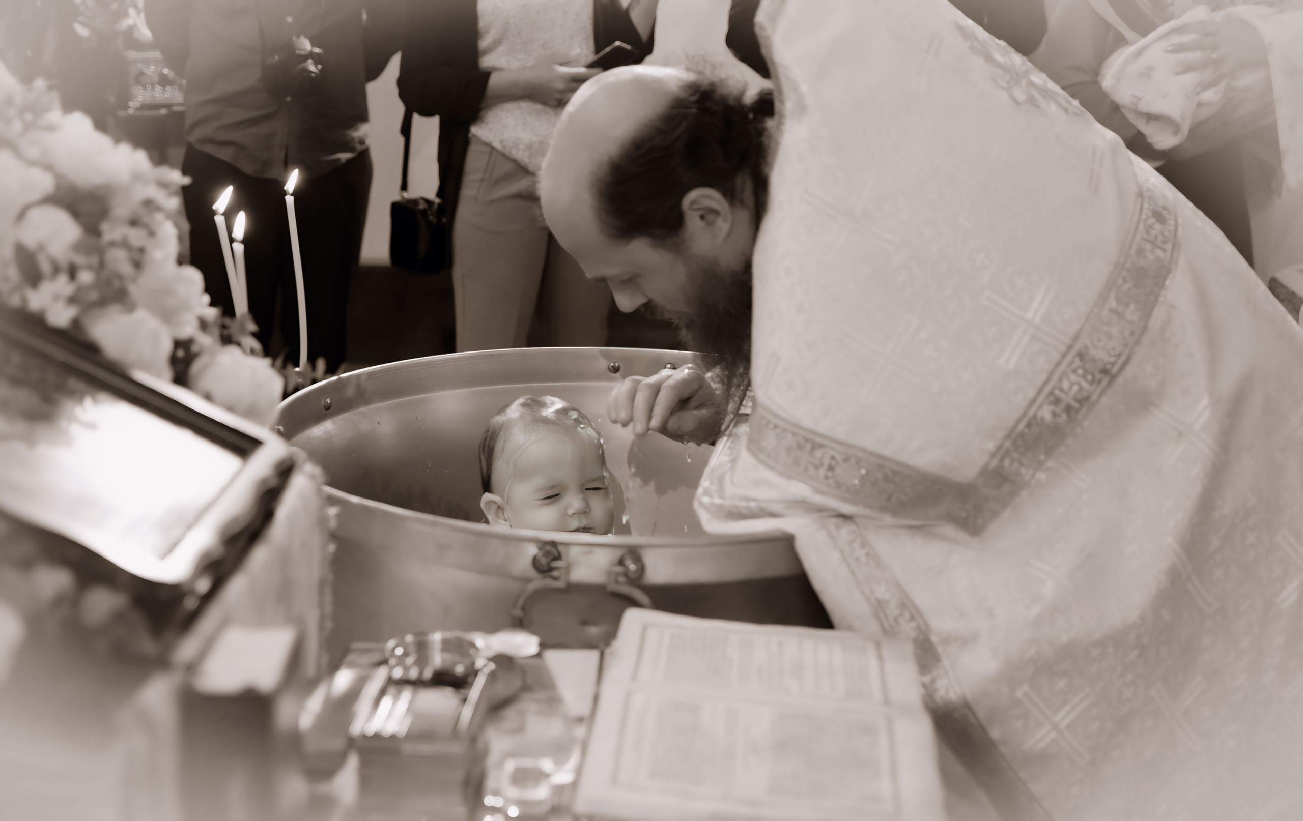 an Orthodox priest puts a baby in a vat and sprinkles it with holy water