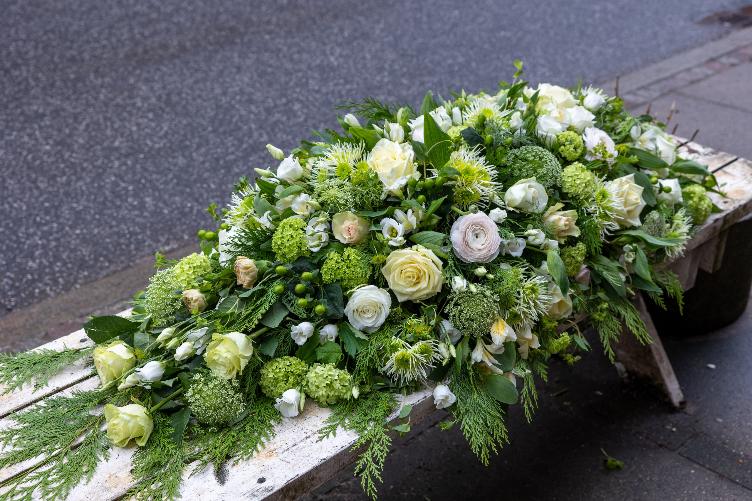Blomsterne er arrangeret tæt på hele kistelåget, som giver en overdådig kiste. De flotte grønne, hvide nuancer er kombineret smukt i sæsonens blomster. Et unikt farvel der tager sig elegant ud i kirken.