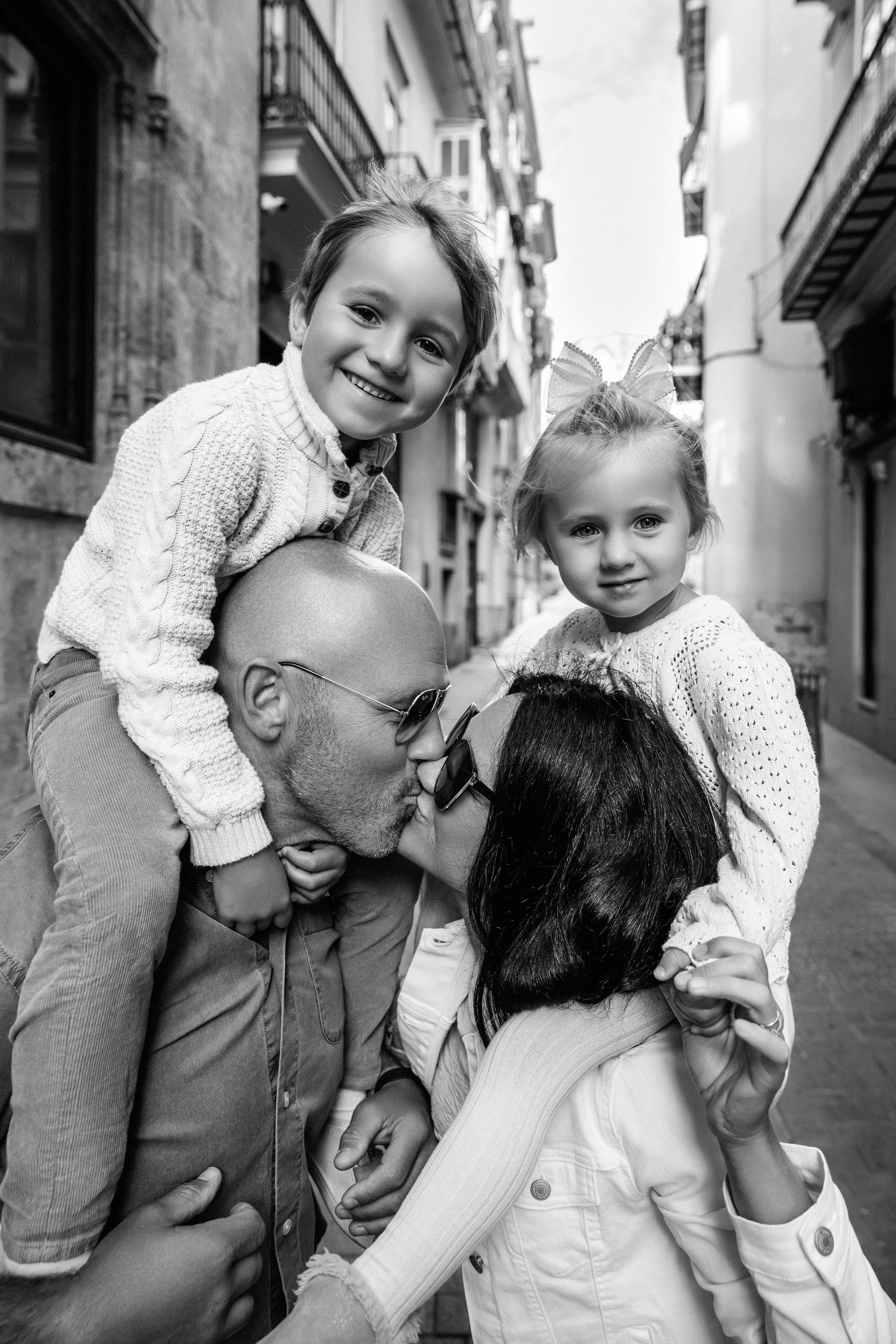 Sesión de fotos familiar en blanco y negro en Valencia, España, con una feliz familia de cuatro compartiendo momentos divertidos con sus hijos por las encantadoras calles del casco histórico de Valencia. Un retrato familiar lifestyle atemporal que transmite sonrisas auténticas, amor y unión familiar. Ideal para fotografía familiar, sesiones de fotos familiares, retratos infantiles, fotografía de vacaciones y fotografía familiar de destino en Valencia y toda España.