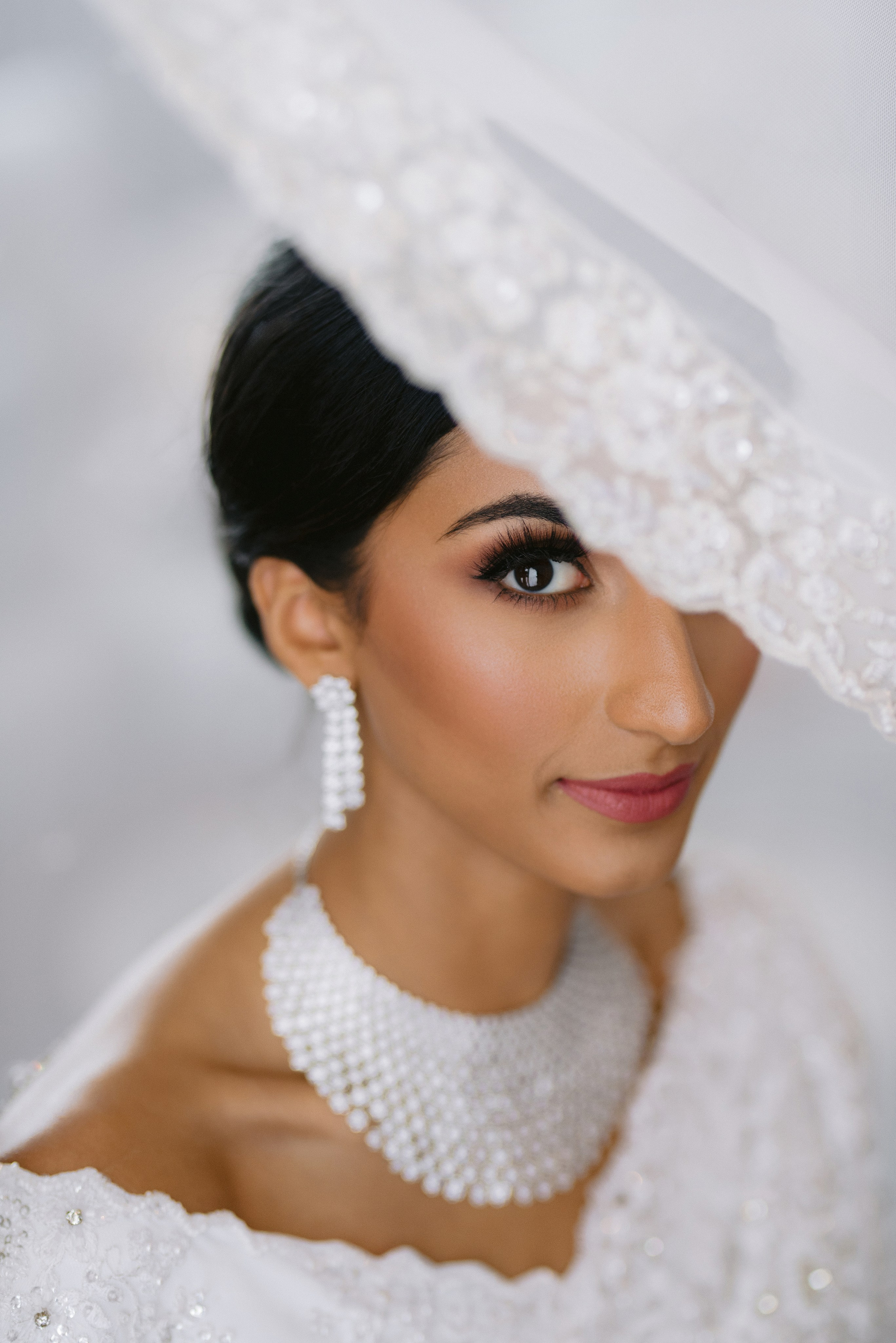 Close-up of a bride with elegant makeup and jewelry, partially covered by her lace veil, looking radiant and confident