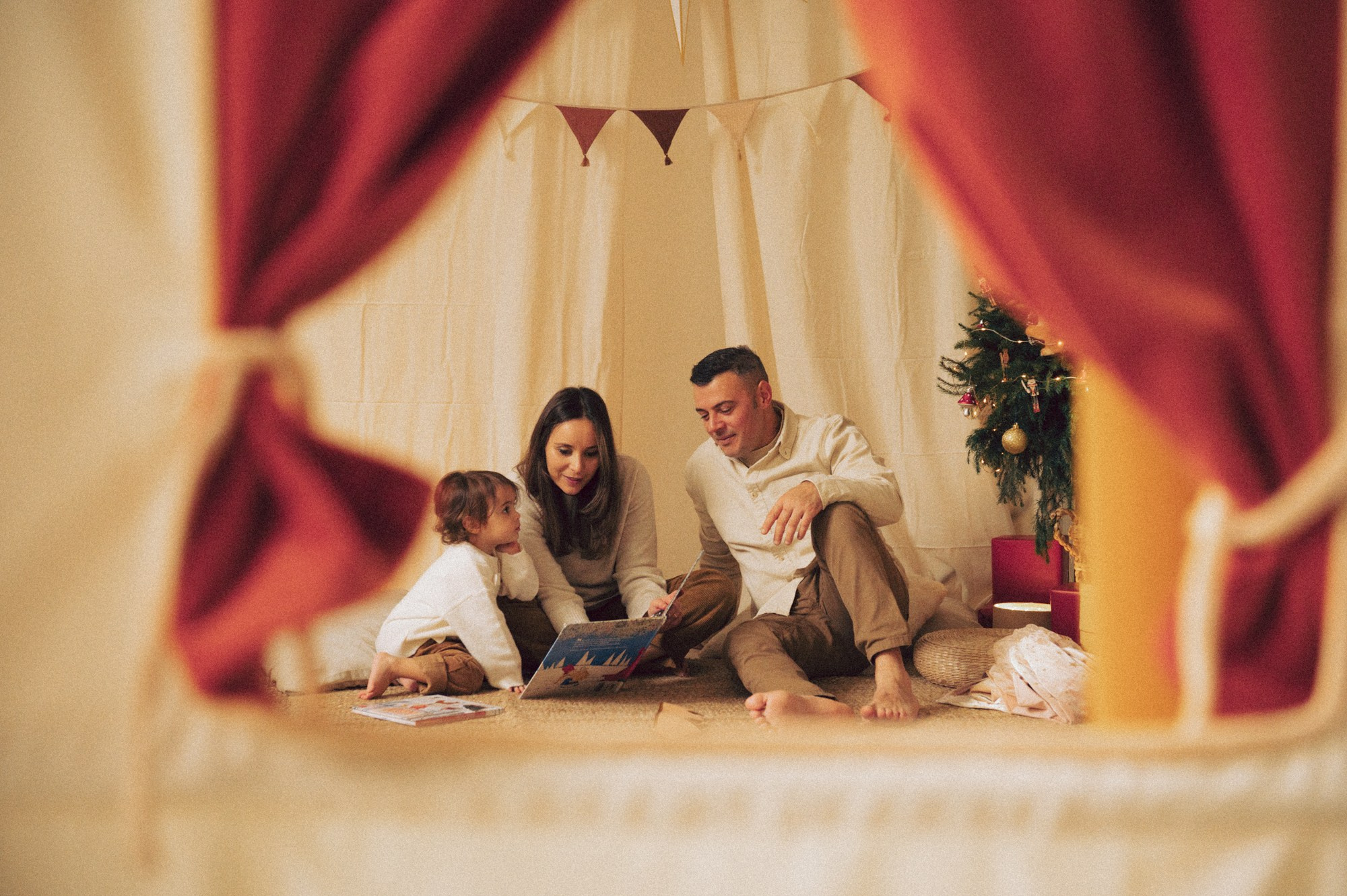 Family Christmas Photoshoot in Milan – Cozy Storytime Inside a Holiday Tent. A tender and intimate moment captured during a Christmas family photoshoot in a natural-light studio in Milan. The family sits together inside a soft holiday tent, reading a Christmas book. The framing through the curtains adds a cinematic, storytelling feel. Ideal for families looking for natural Christmas portraits, warm and emotional family photography, and cozy studio holiday sessions. Family Christmas photoshoot Milan, natural family portraits, holiday studio session, Christmas storytelling photography, kids and family Christmas photos. Family reading a Christmas book inside a holiday tent, viewed through red curtains.
