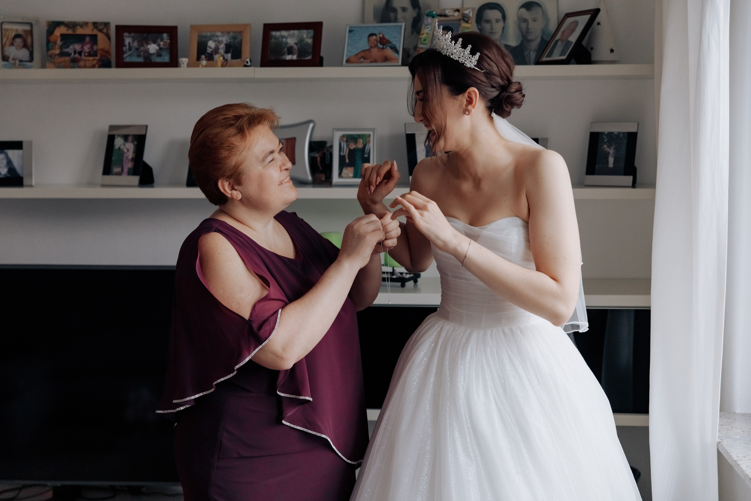 Die Mutter hilft der Braut, die Schmuckstücke anzulegen, während sie beide lachen. Ein herzlicher und fröhlicher Moment der Hochzeitsvorbereitung.