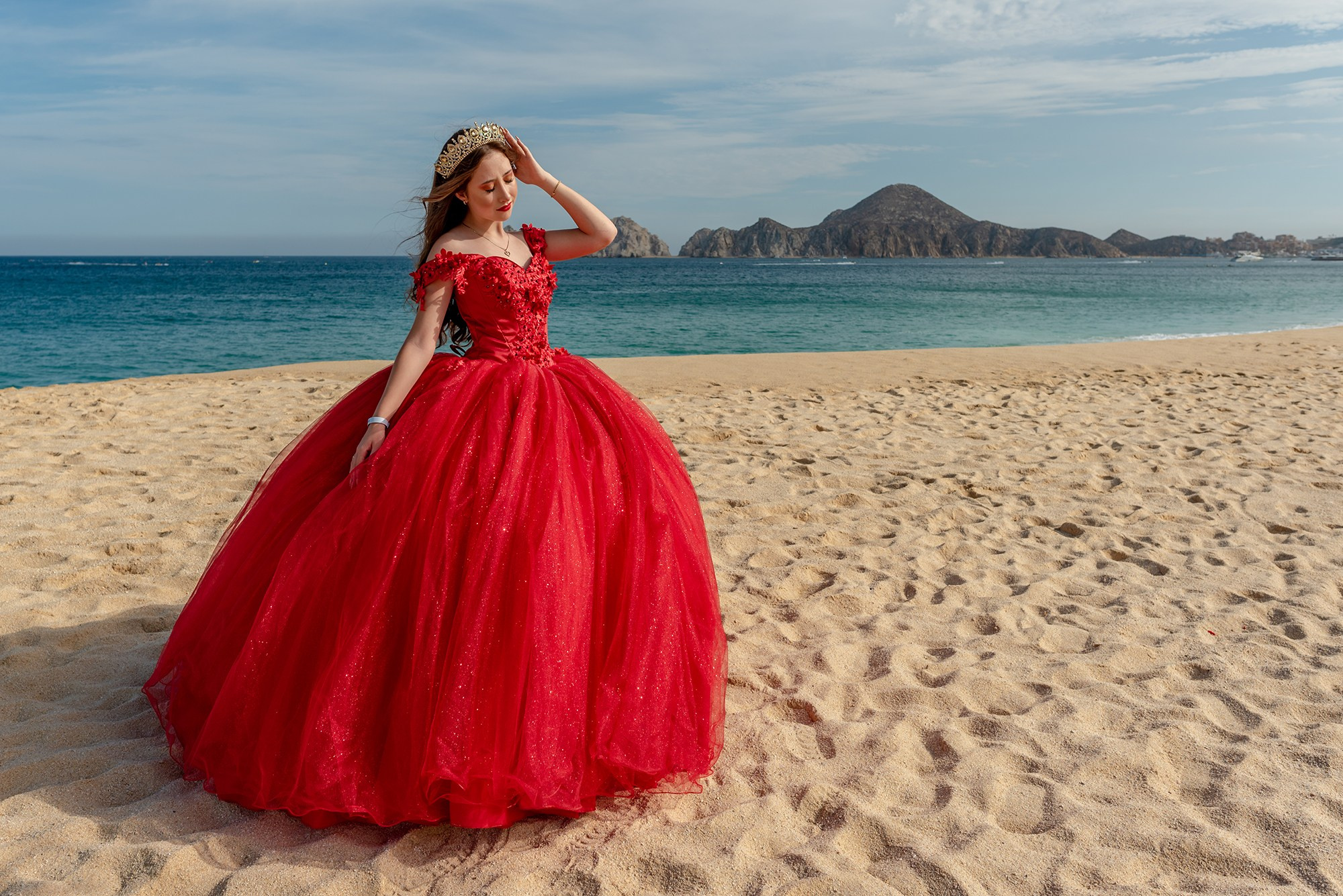 Los Cabos quinceañera photoshoot – Sofie’s XV años session at El Médano beach in Cabo San Lucas, featuring a red quinceañera gown, roses bouquet, and the iconic Finisterra rocky cliffs as backdrop