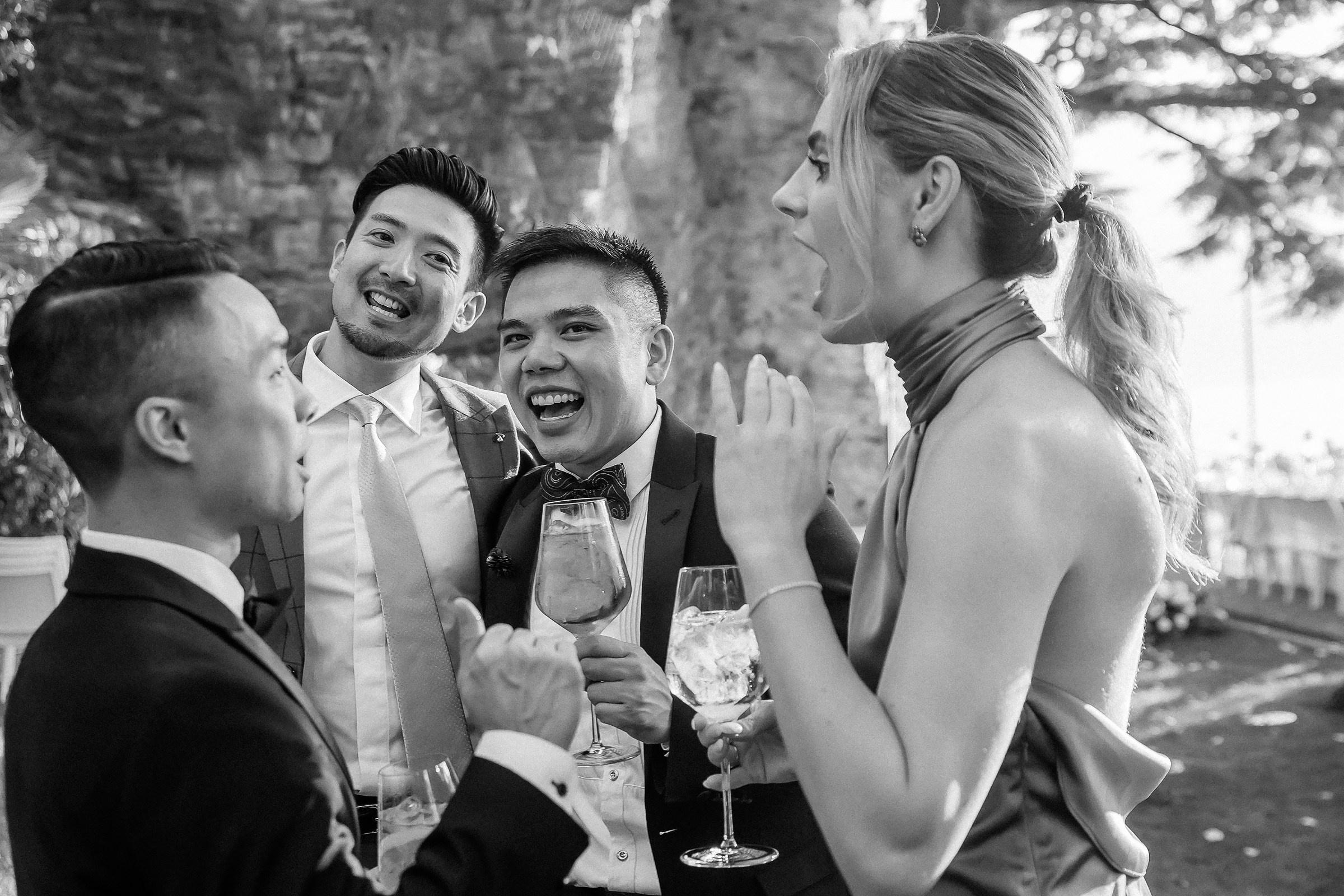 Four guests in formal attire share laughter and conversation, holding wine glasses in a lively outdoor setting.