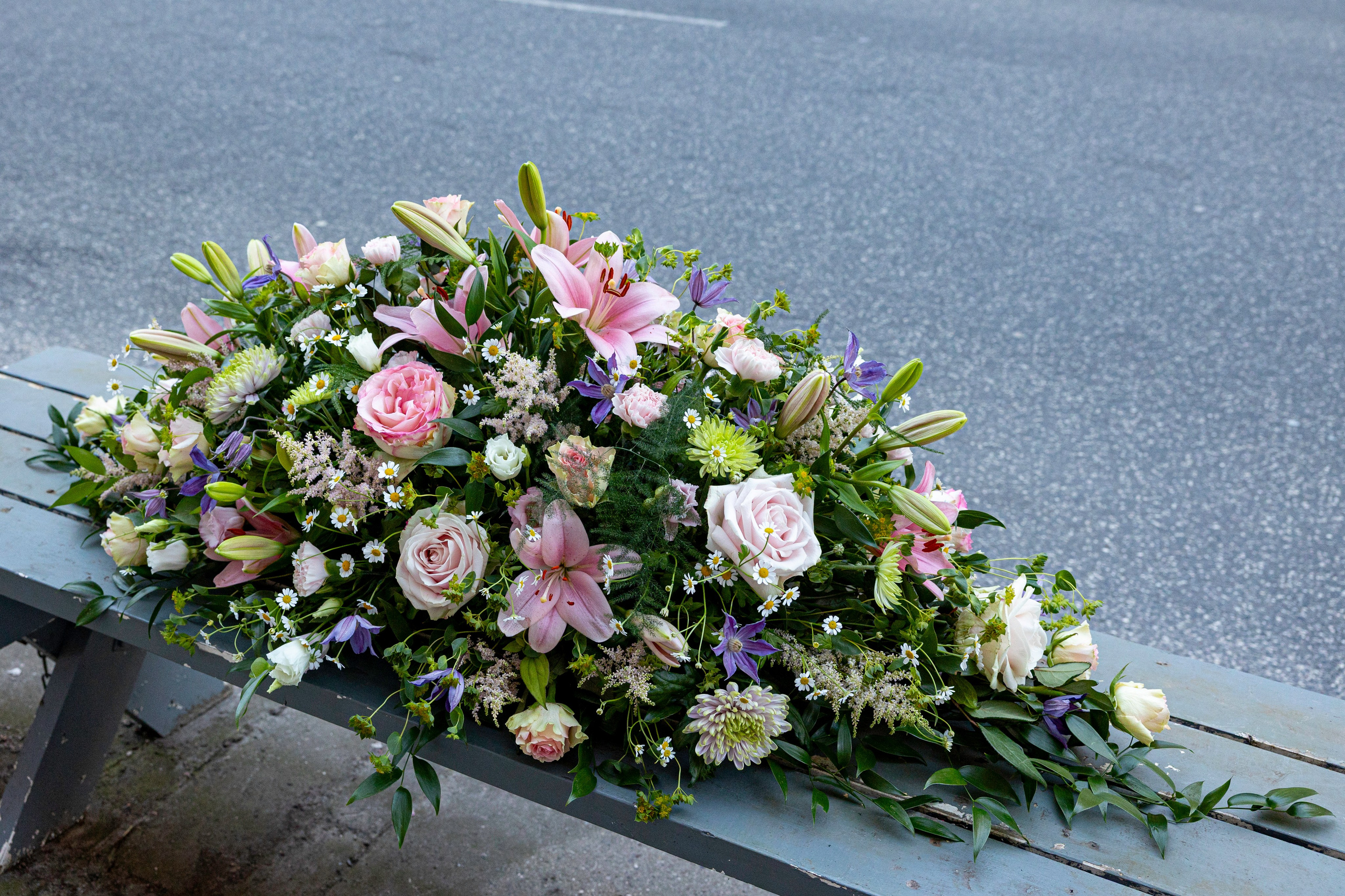 Elegant kistepynt med lyserøde liljer, roser og fine sæsonblomster. Håndlavet af Akropolis Blomster som en smuk og værdig sidste hilsen, der skaber en rolig og respektfuld ramme om afskeden.