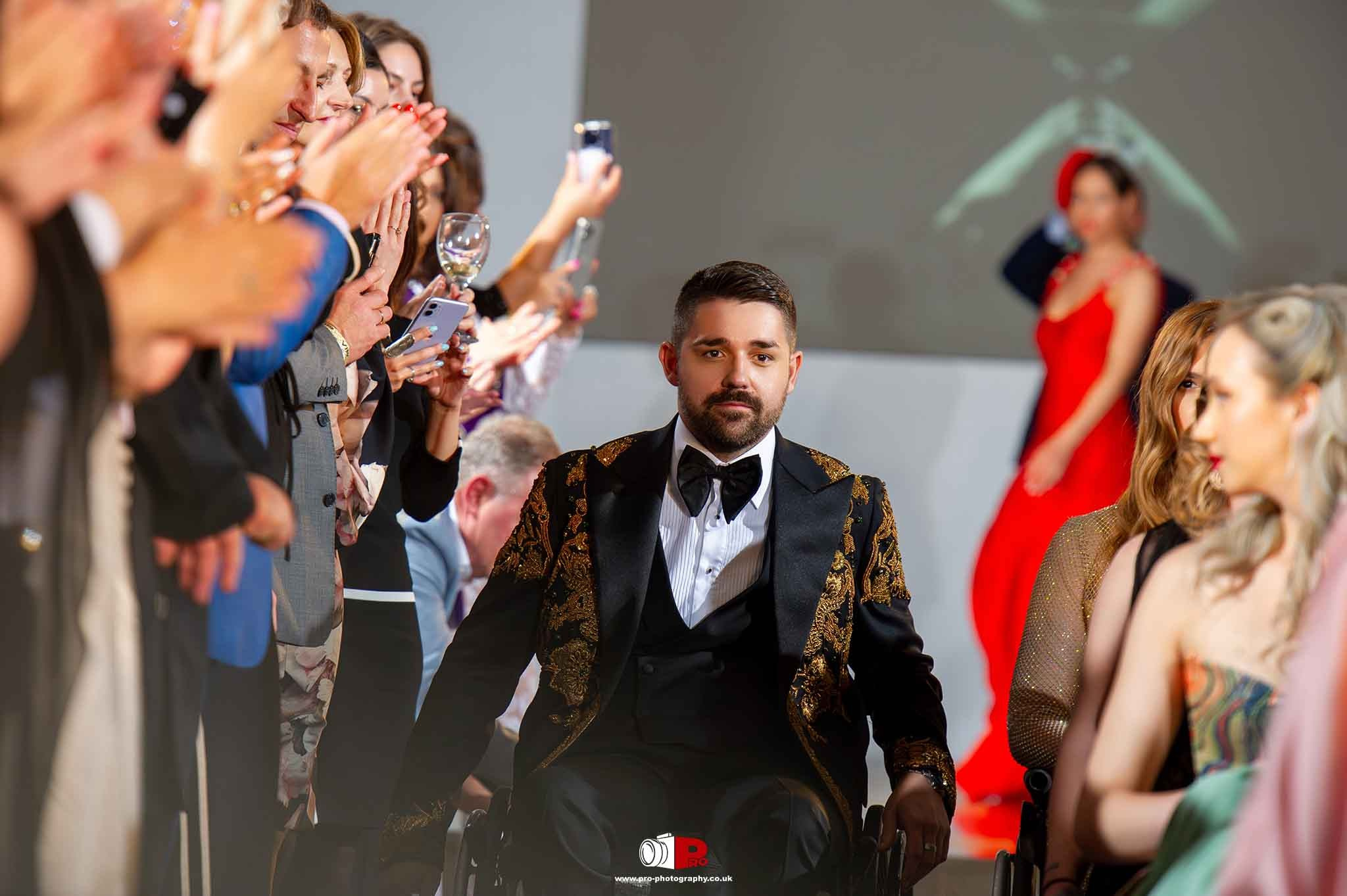 A confident model in a wheelchair dressed in an ornate gold and black suit receiving applause at a fashion event.
