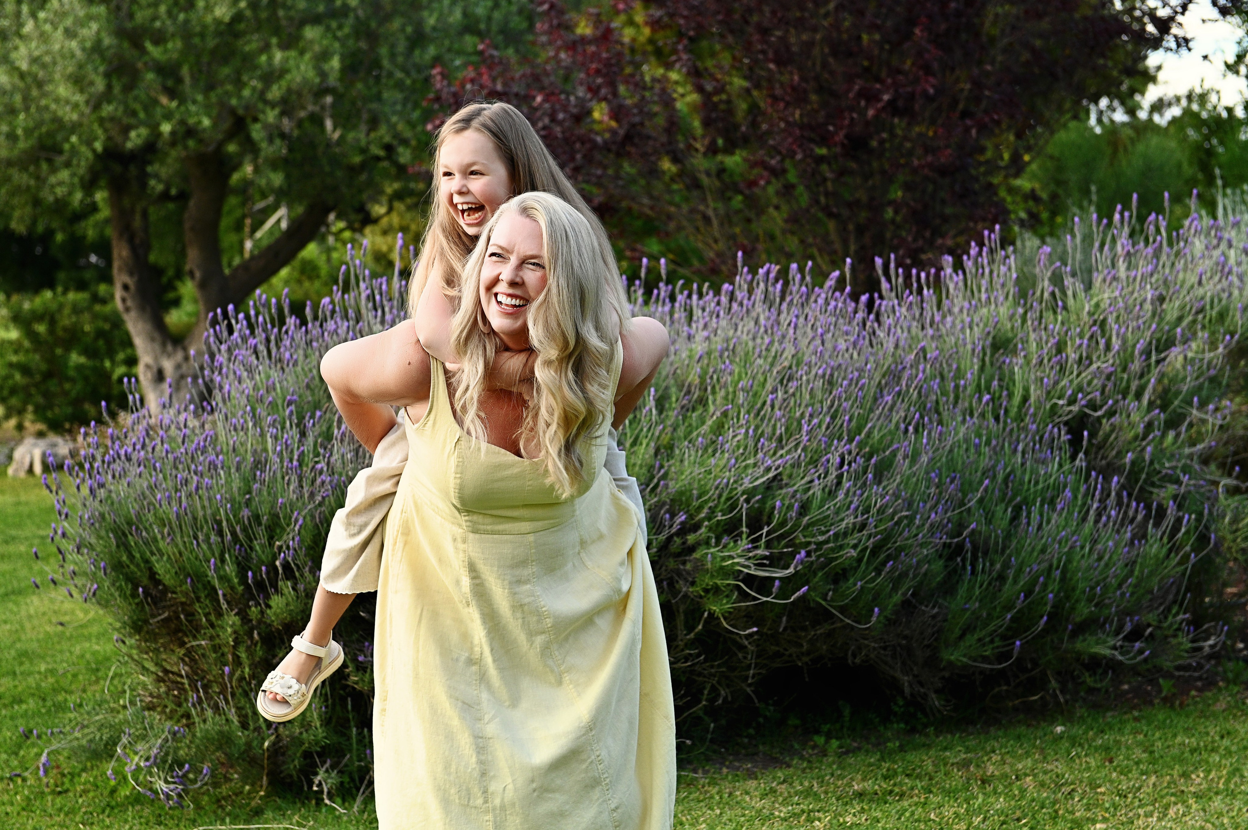 Servizio fotografico di famiglia in Puglia: una mamma in abito giallo estivo tiene la figlia sulle spalle mentre ridono vicino a cespugli di lavanda.
