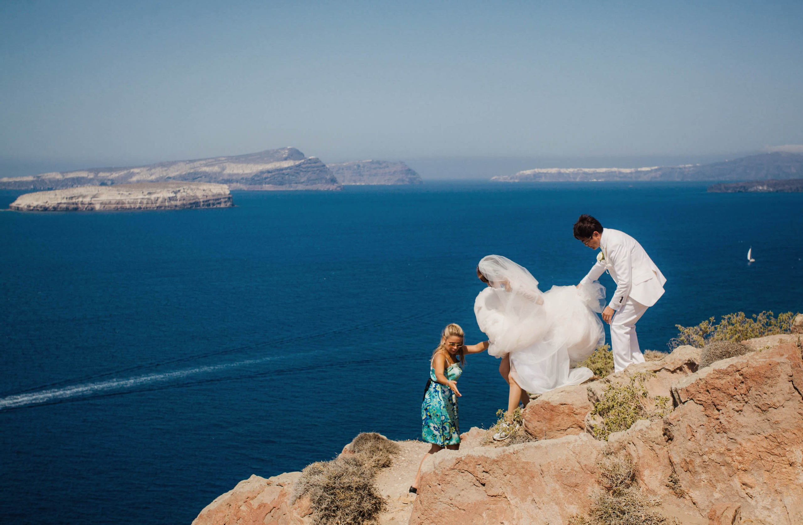 A bride is assisted by a woman and her groom as they scale the cliffs of Santorini in pursuit of an epic location for a wedding portrait.