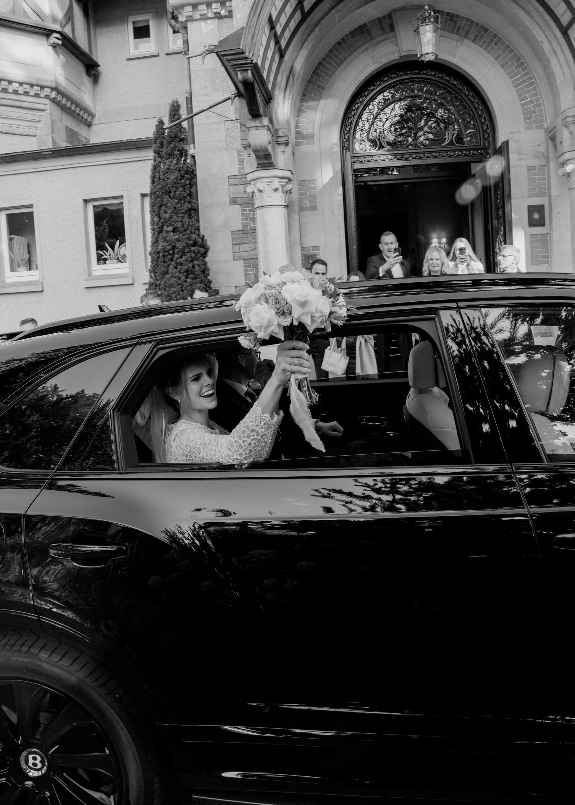 Bride holding white bouquet from inside black Bentley at church entrance, wedding Frankfurt