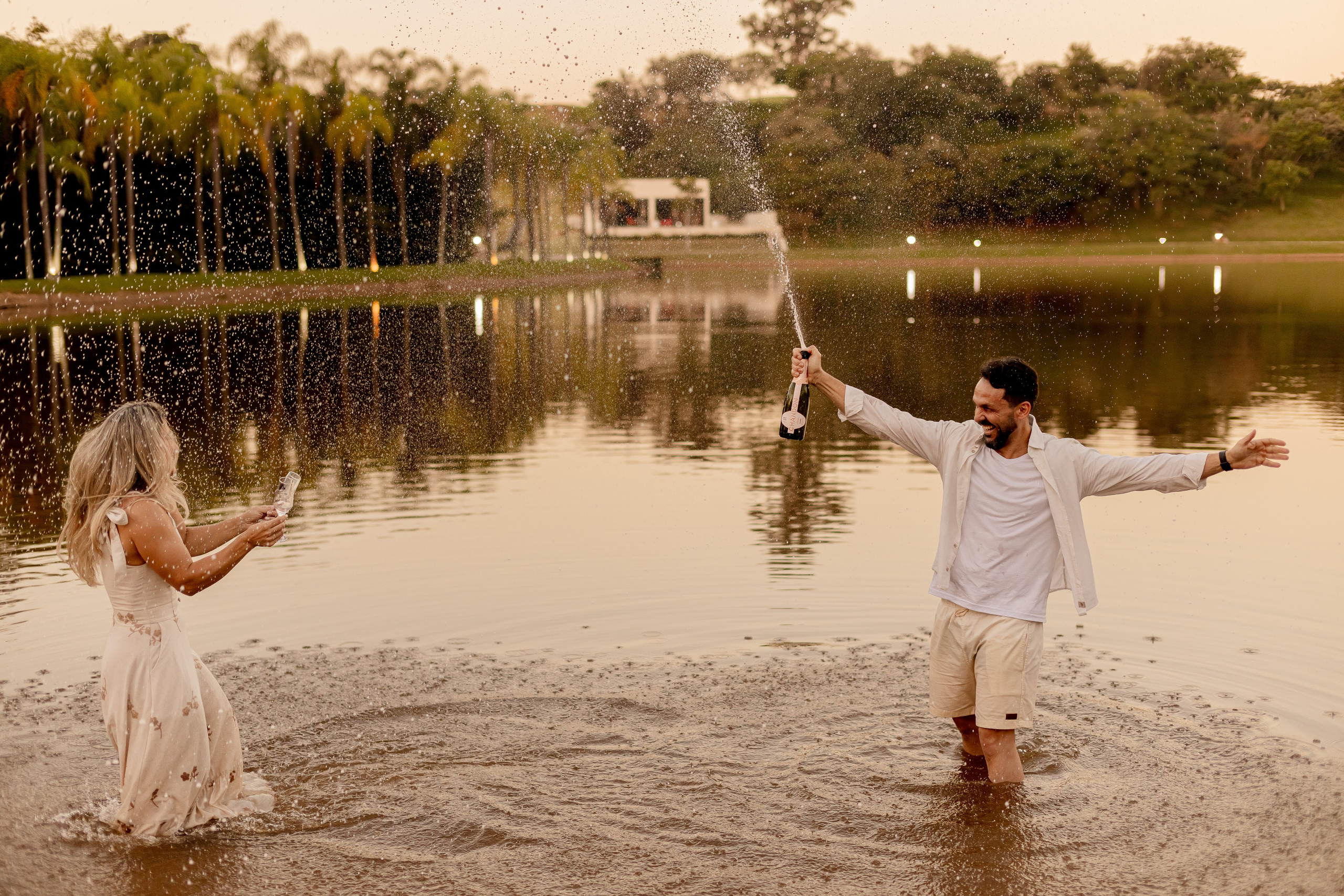 Casal celebrando com champanhe dentro do lago no Resort das Oliveiras ao entardecer, com espirros d'água e sorrisos