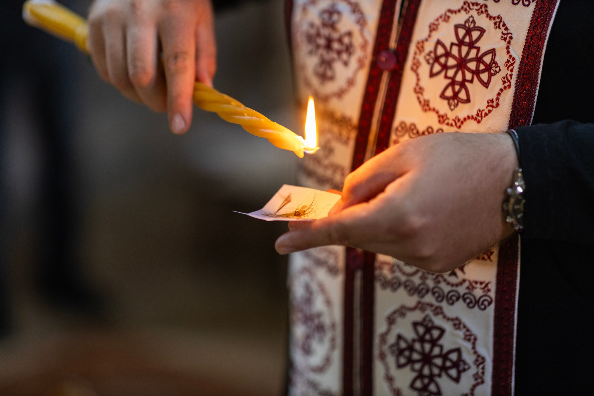 Krupni plan sveštenikovih ruku koje drže upaljenu, uvrnutu žutu sveću iznad male papirne sličice tokom religijskog obreda, sa prikazom šarene (ornamentisane) odore.