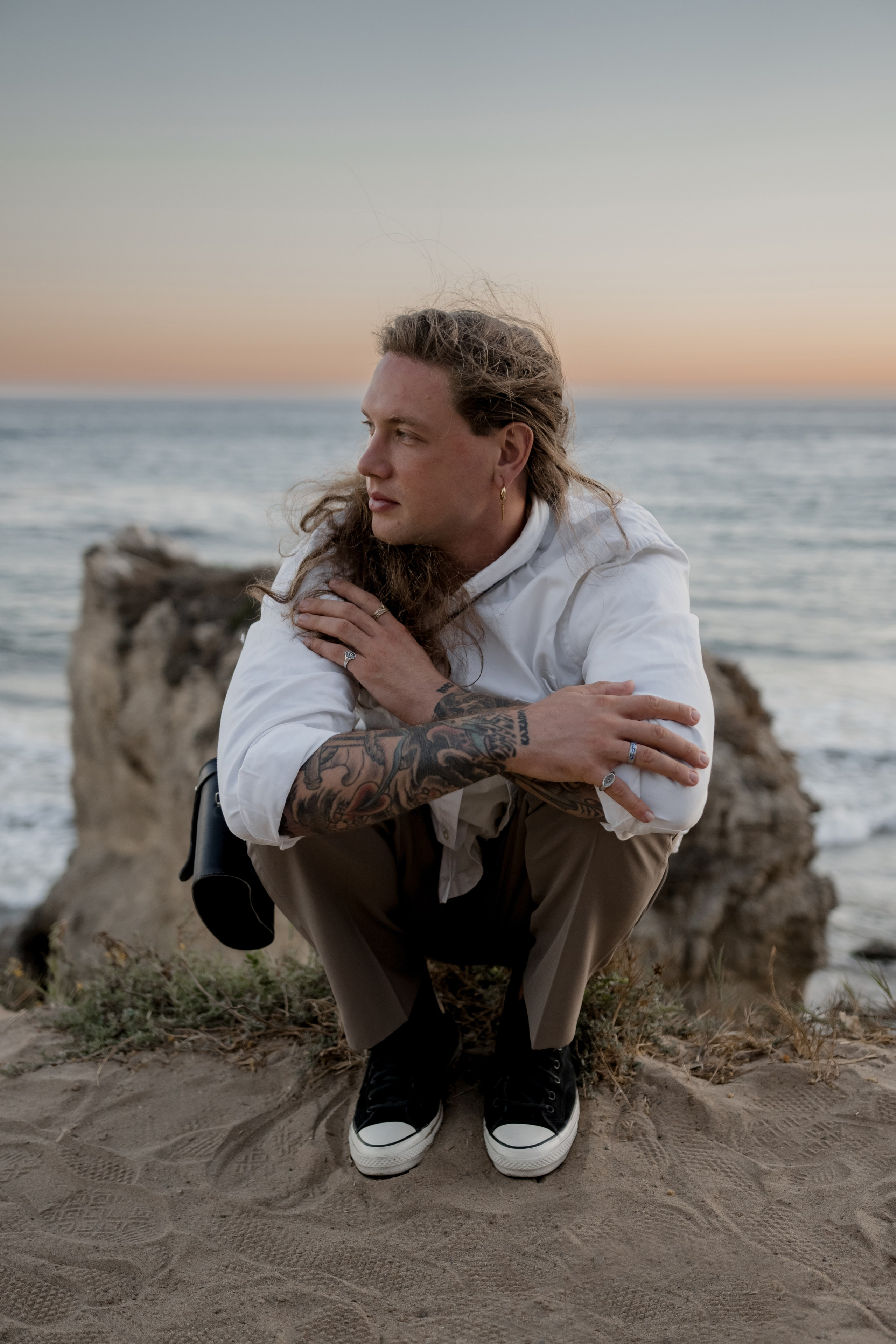 Tattooed male model crouching near the beach, emphasizing modern urban fashion in a natural setting.