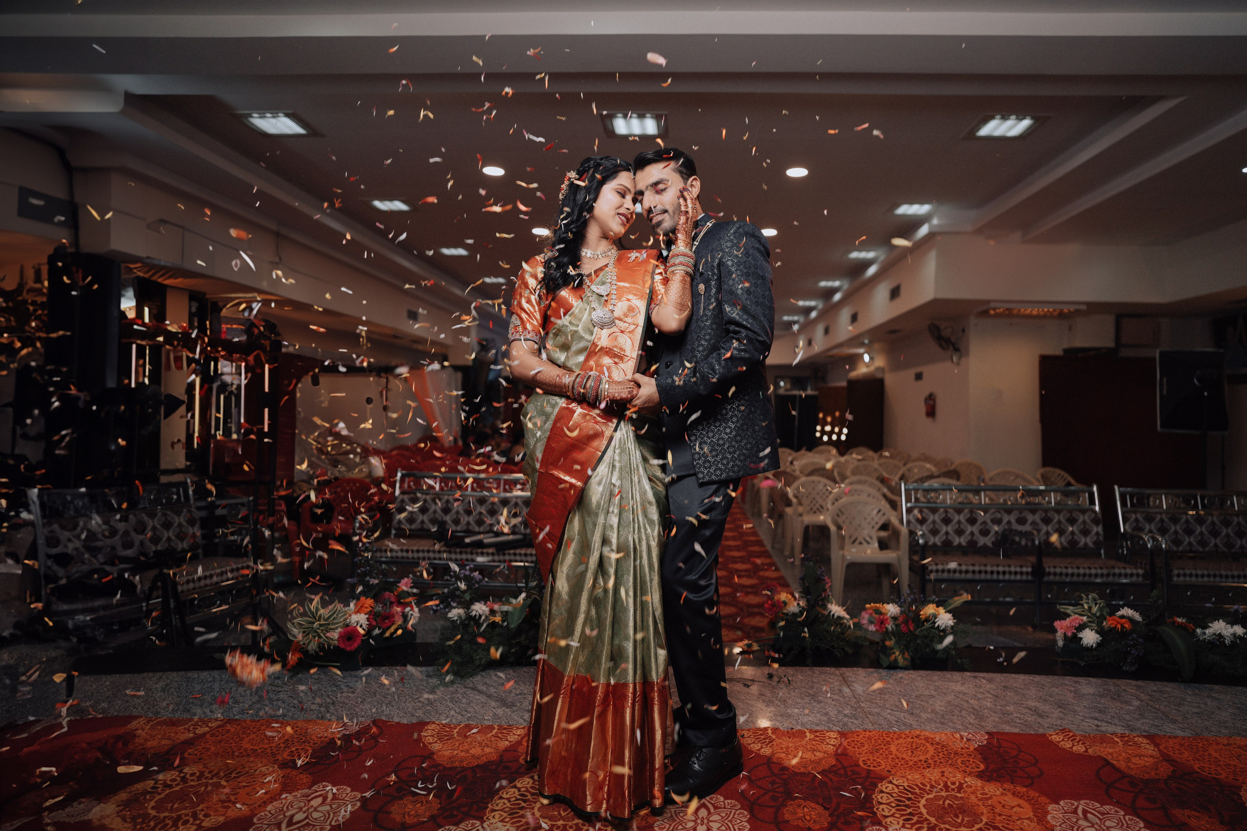 candid indoor photo of a newly married South Indian couple embracing with confetti falling in a wedding hall in Malleshwaram, Bengaluru