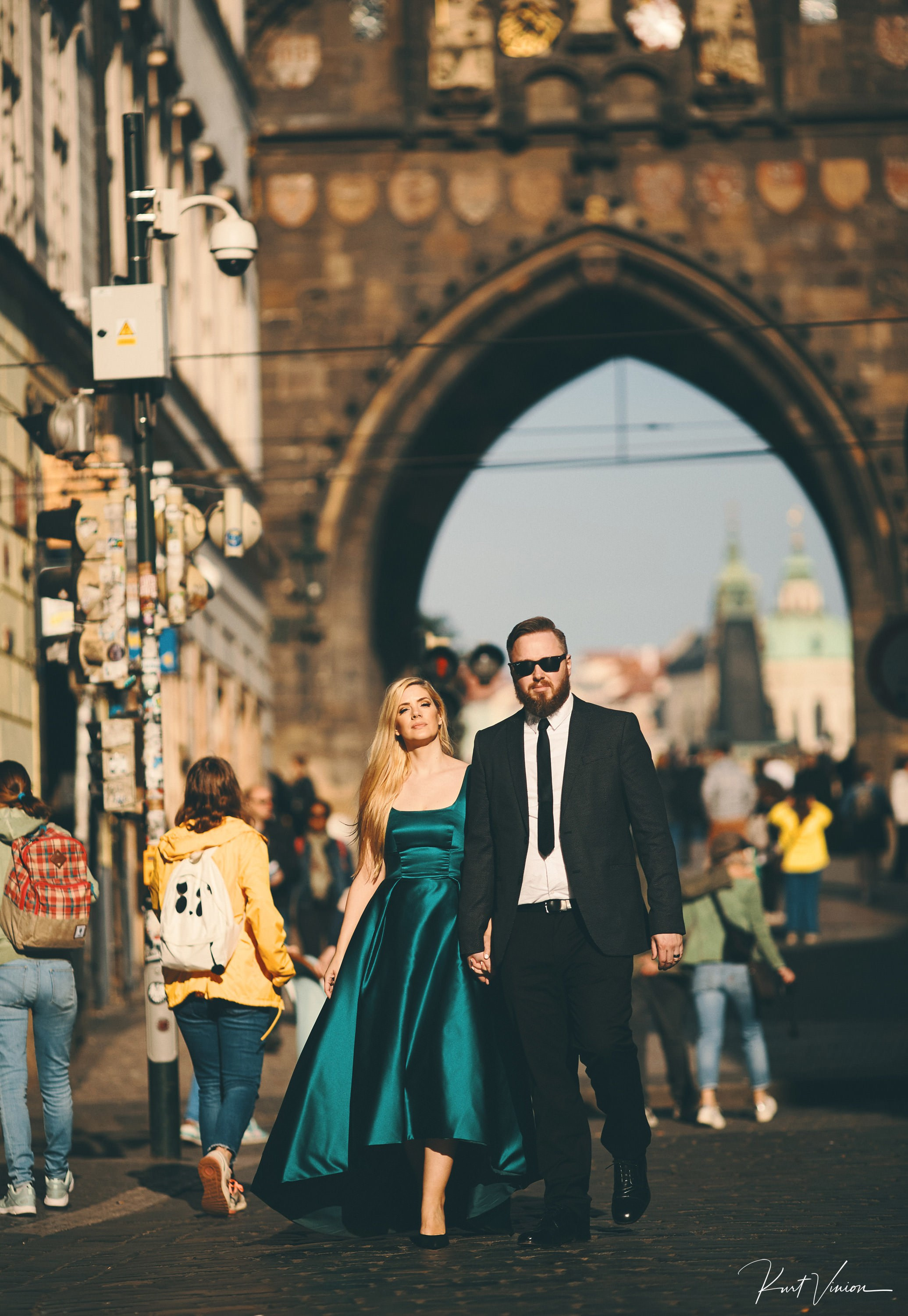 Blonde-haired woman in a silk dark green dress walks confidently hand-in-hand with her bearded suit wearing partner as the gothic tower of the Charles Bridge towers above them
