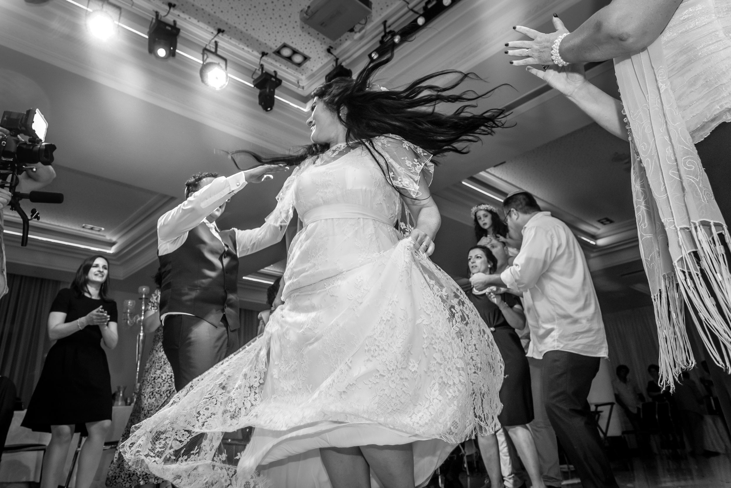 The bride dancing energetically in a lace wedding dress with flowing hair, surrounded by guests clapping and watching. The banquet hall's decor and ceiling lighting are visible in the background.