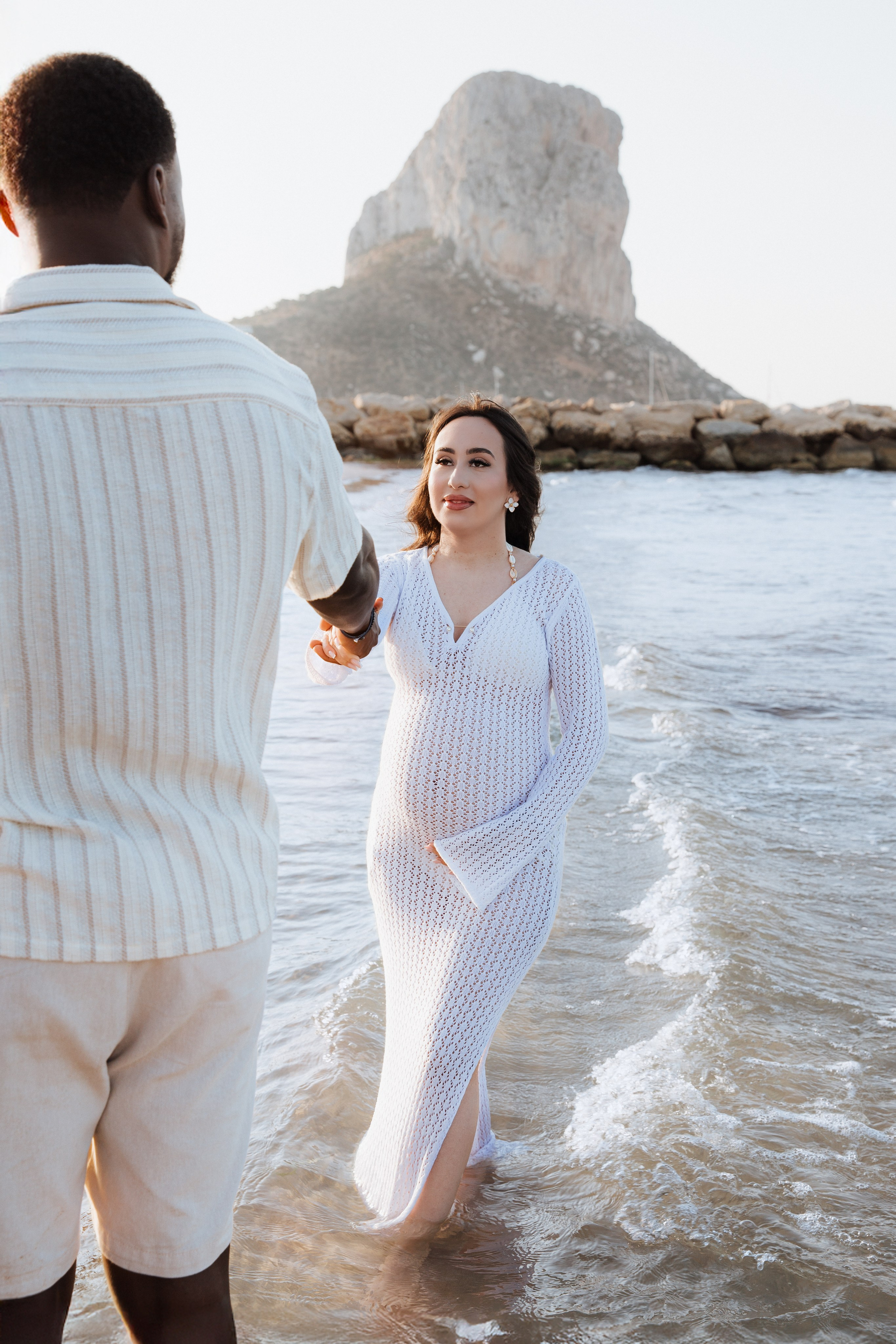 Sesión romántica de embarazo en Calpe, España — mujer embarazada con vestido blanco caminando por la orilla, tomada de la mano con su pareja con el Peñón de Ifach al fondo. Una sesión serena y emocional ideal para quienes buscan fotos de maternidad en Calpe y la costa española.