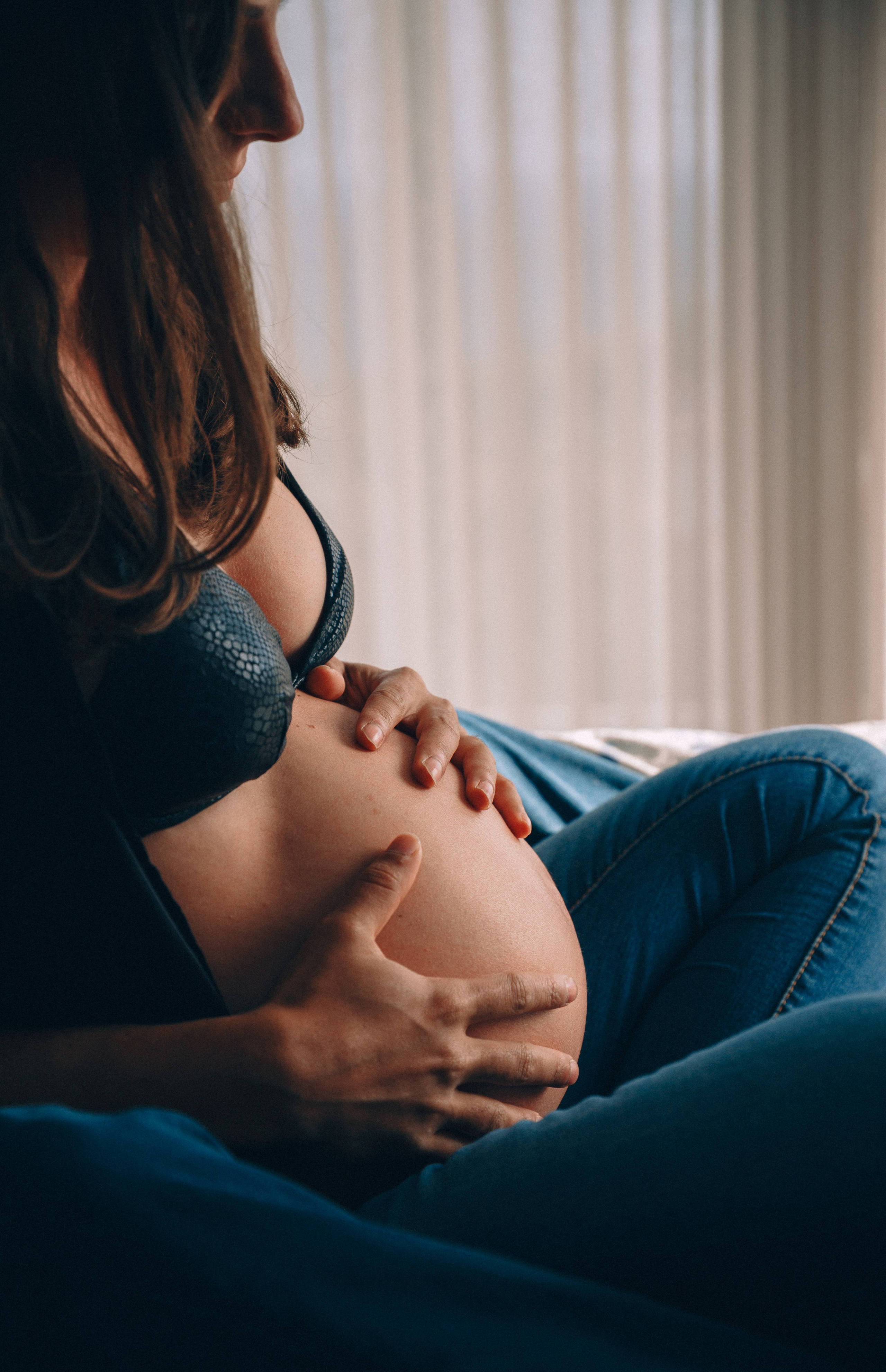 Retrato íntimo de maternidad de una mujer con las manos sobre su vientre descubierto, bañada por luz natural cerca de cortinas translúcidas — momento de calma capturado en un hogar acogedor en Valencia, España.