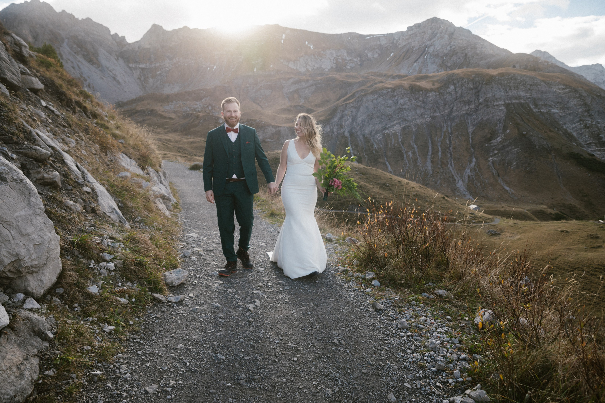 Brautpaar geht Bergpfad entlang, sonnenbeschienene Gipfel im Hintergrund