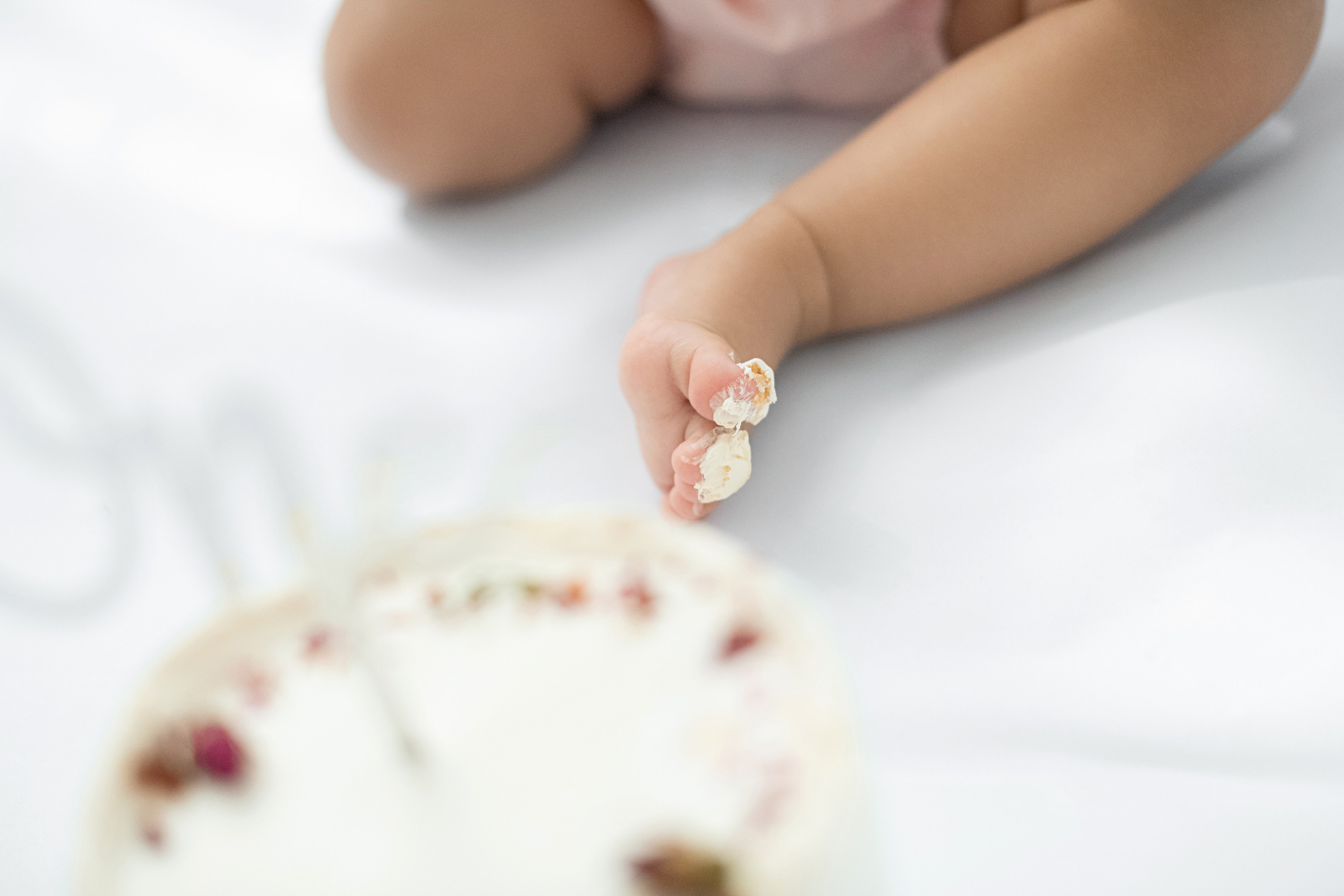 Baby’s chubby toes pinching frosting next to a white cake; blurred legs in background on white cloth.
