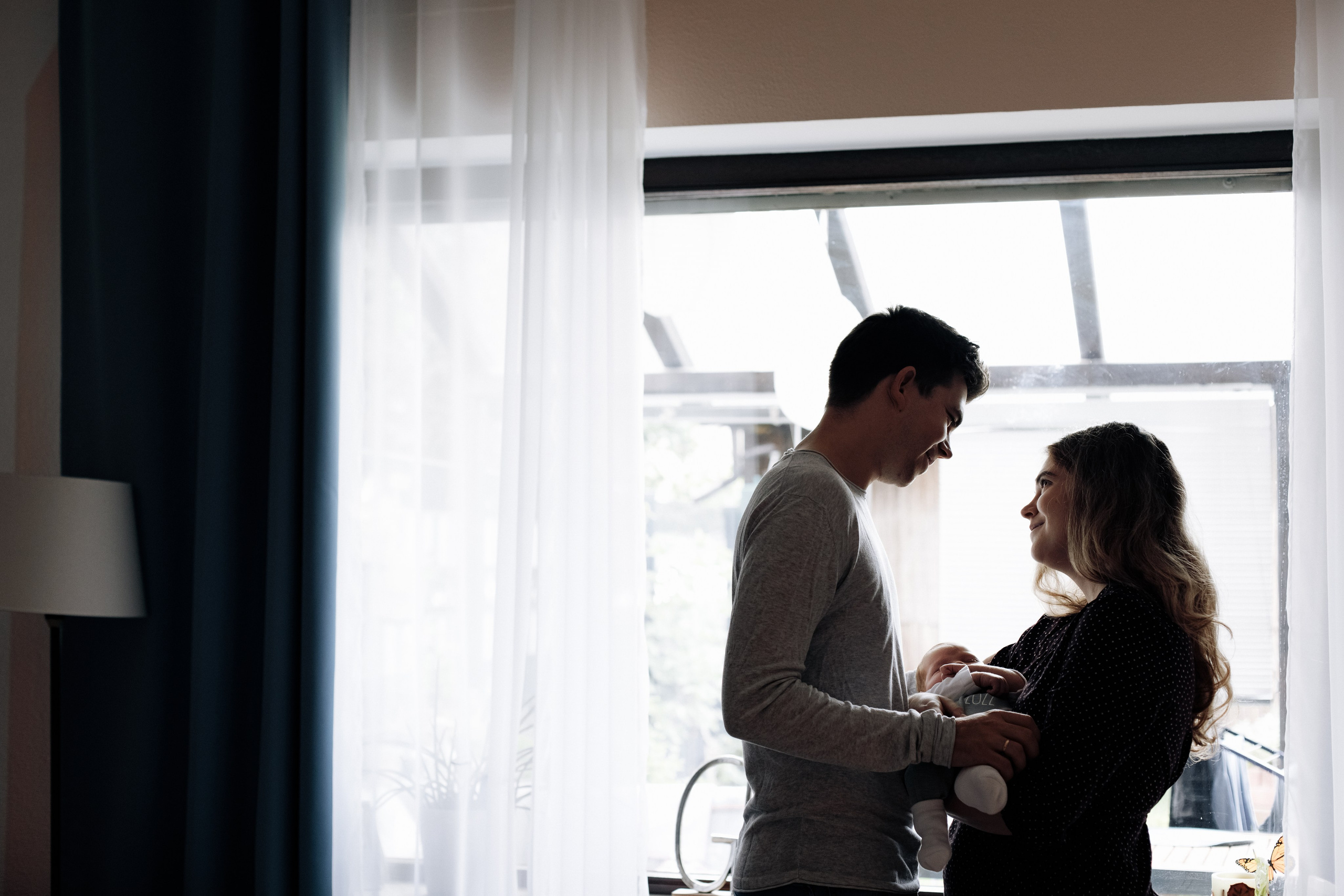 Vater und Mutter stehen am Fenster, halten ihr Baby im Arm und schauen sich liebevoll an.