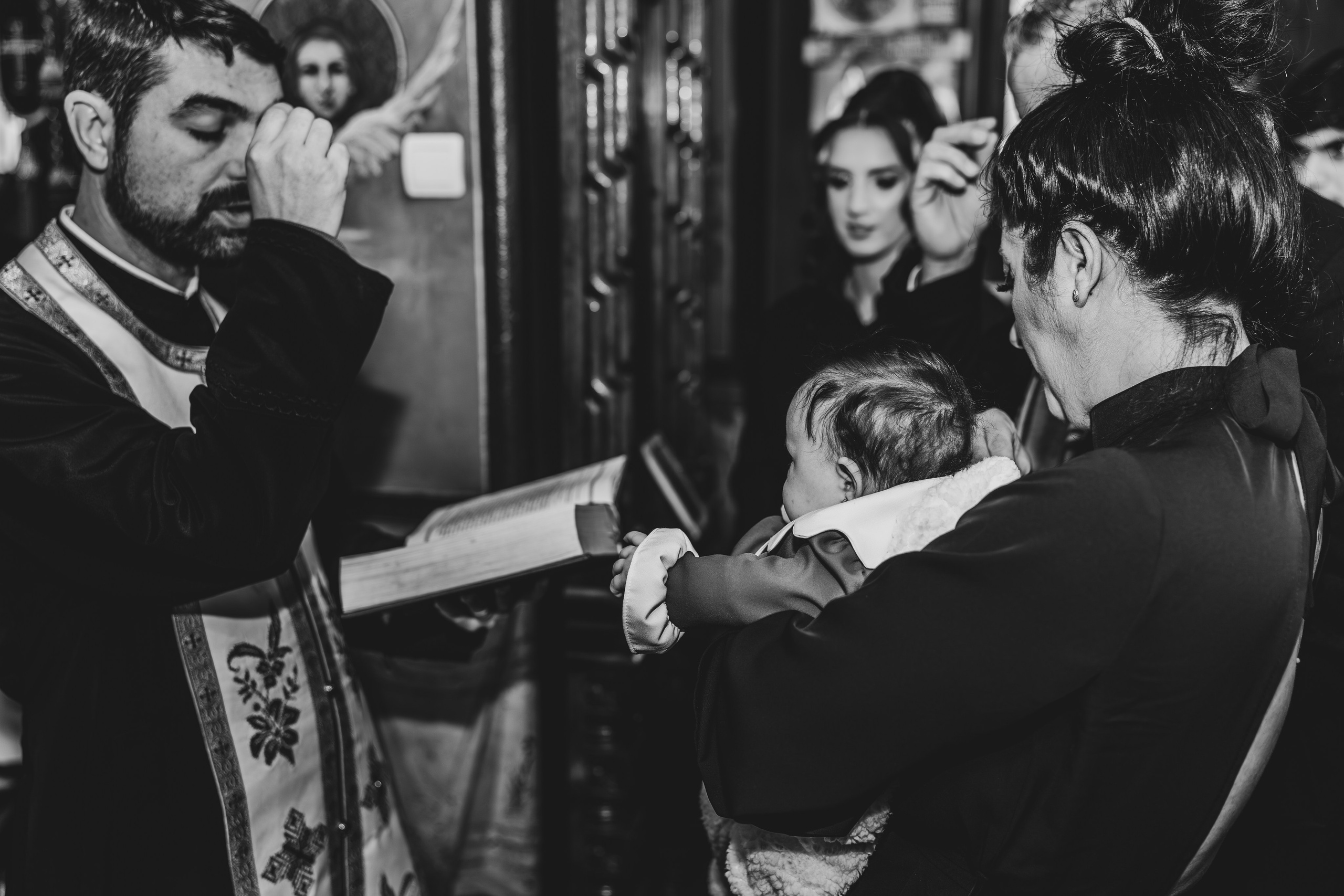 Cadru alb-negru de ansamblu de la ceremonia de botez a Evelinei Maria în Biserica Grivița. Preotul, nașii și familia apropiată sunt uniți în momentul central al binecuvântării. Fotografie solemnă și plină de emoție.