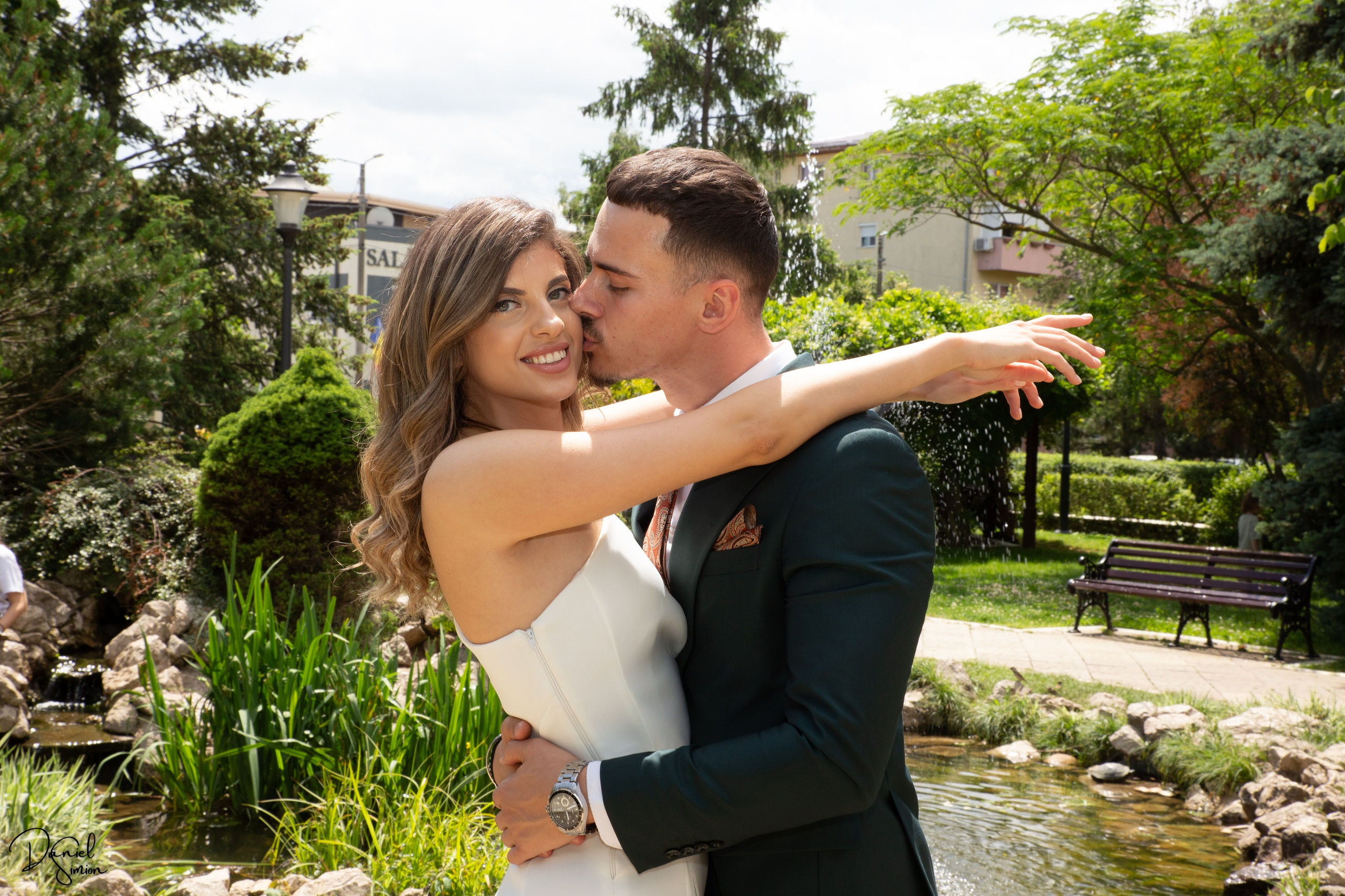 Couple embracing near a pond in a garden with trees and greenery around.
