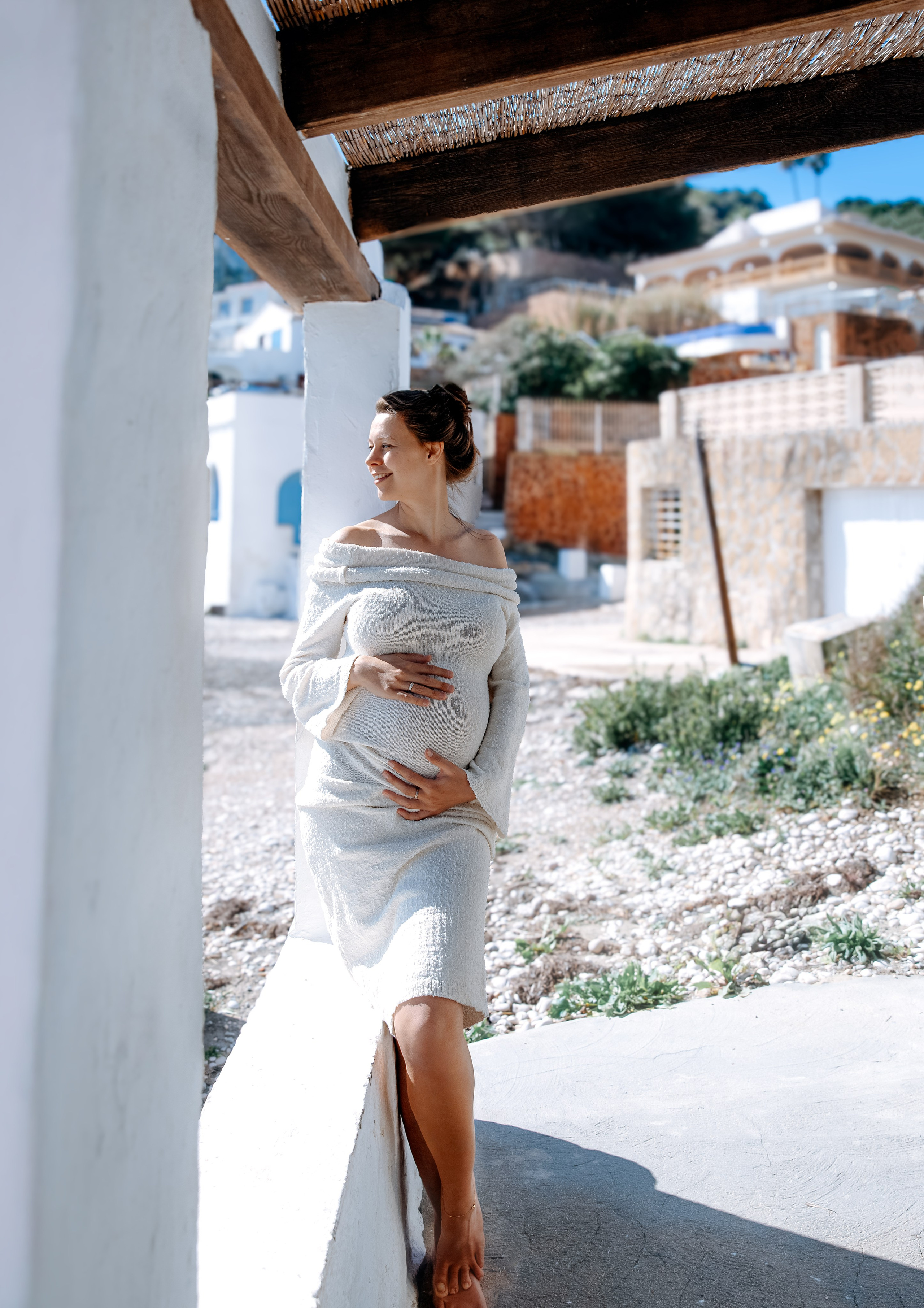Retrato radiante de embarazo en Valencia, España — mujer con vestido crema de hombros descubiertos apoyada en una columna blanca en un pueblo costero de estilo mediterráneo bajo la luz del sol.